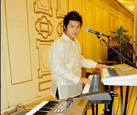 Man playing keyboard at an event, wearing a cream-colored shirt, with a microphone. Gold-colored walls in the background.