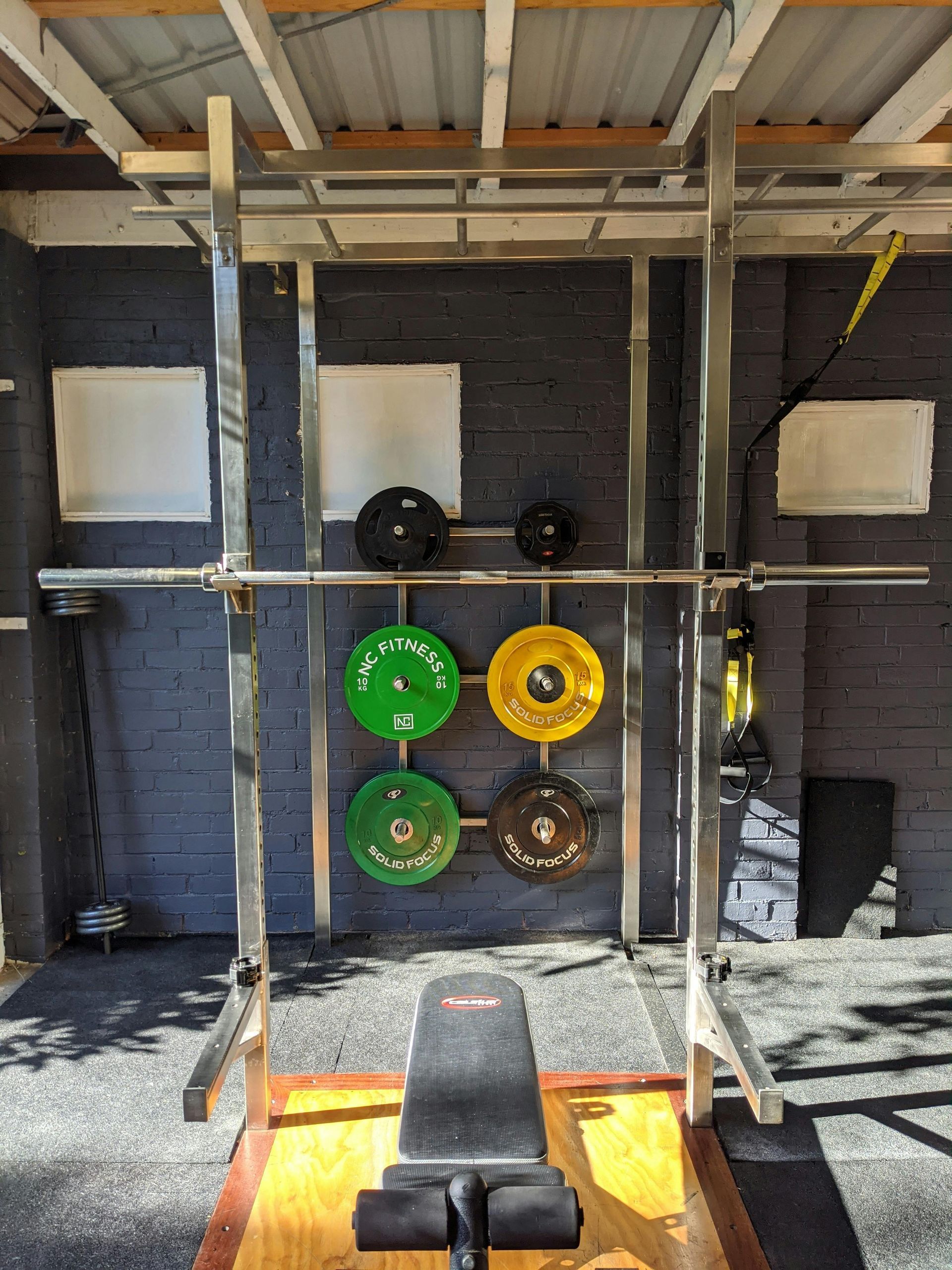 Weightlifting rack with barbell, weight plates, and a bench. Green, yellow, and brown plates. Black wall backdrop.
