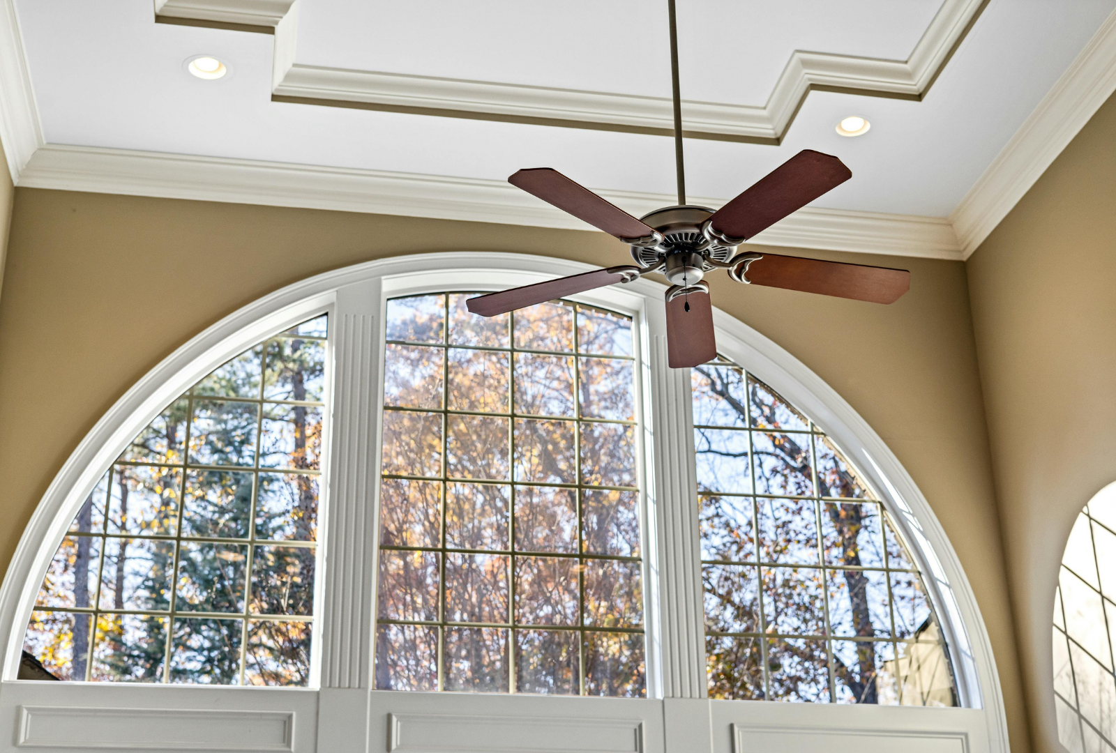 A ceiling fan hangs in front of a large, arched window with a grid pattern, set against tan walls and a tray ceiling.