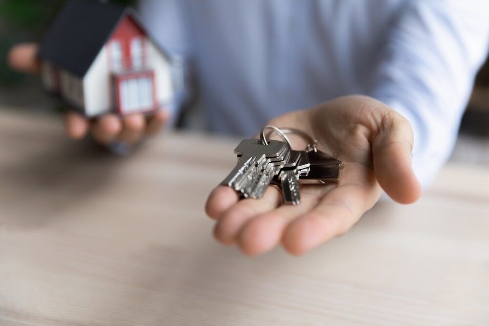 A Person is Holding a Bunch of Keys in Their Hand and a Small House in the Background — Key2 Conveyancing In Tuncurry, NSW