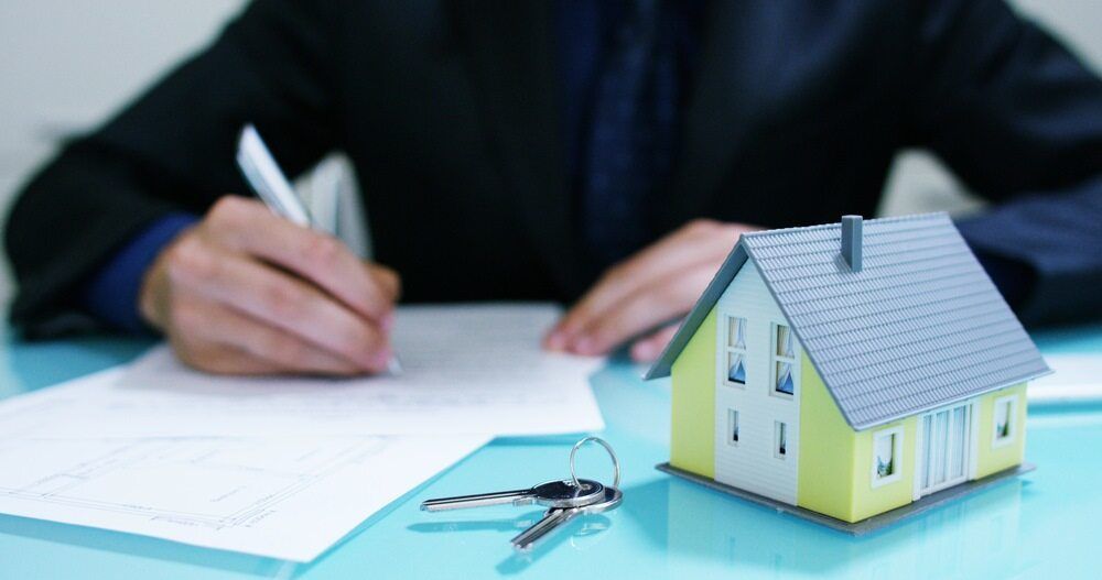 A Man is Writing on a Piece of Paper Next to a Model House and Keys — Key2 Conveyancing In Pacific Palms, NSW