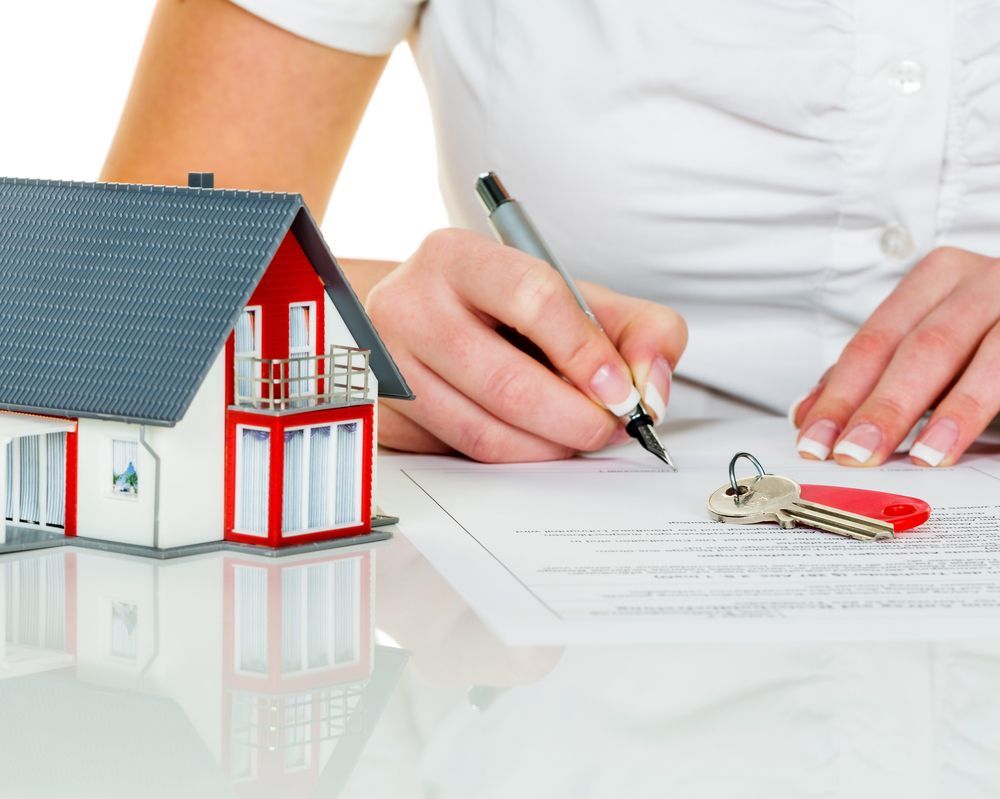 A Woman is Writing on a Piece of Paper Next to a Model House and Keys — Key2 Conveyancing In Old Bar, NSW