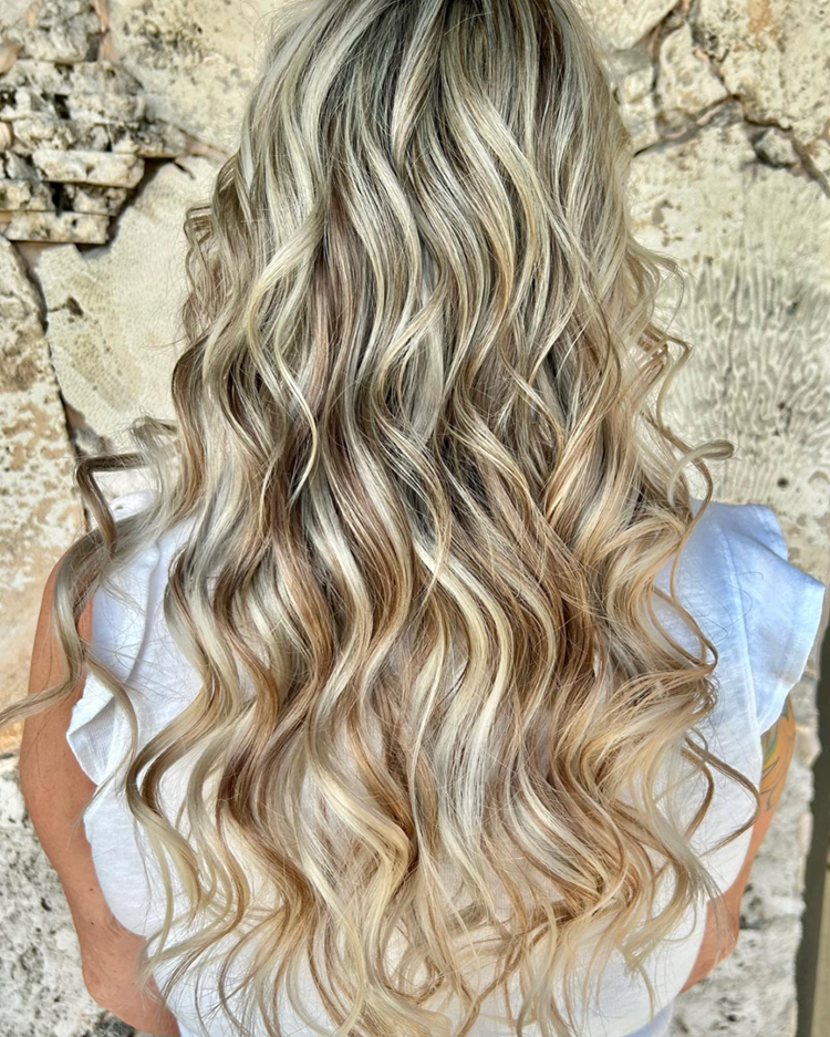 A woman with long blonde wavy hair is standing in front of a stone wall.