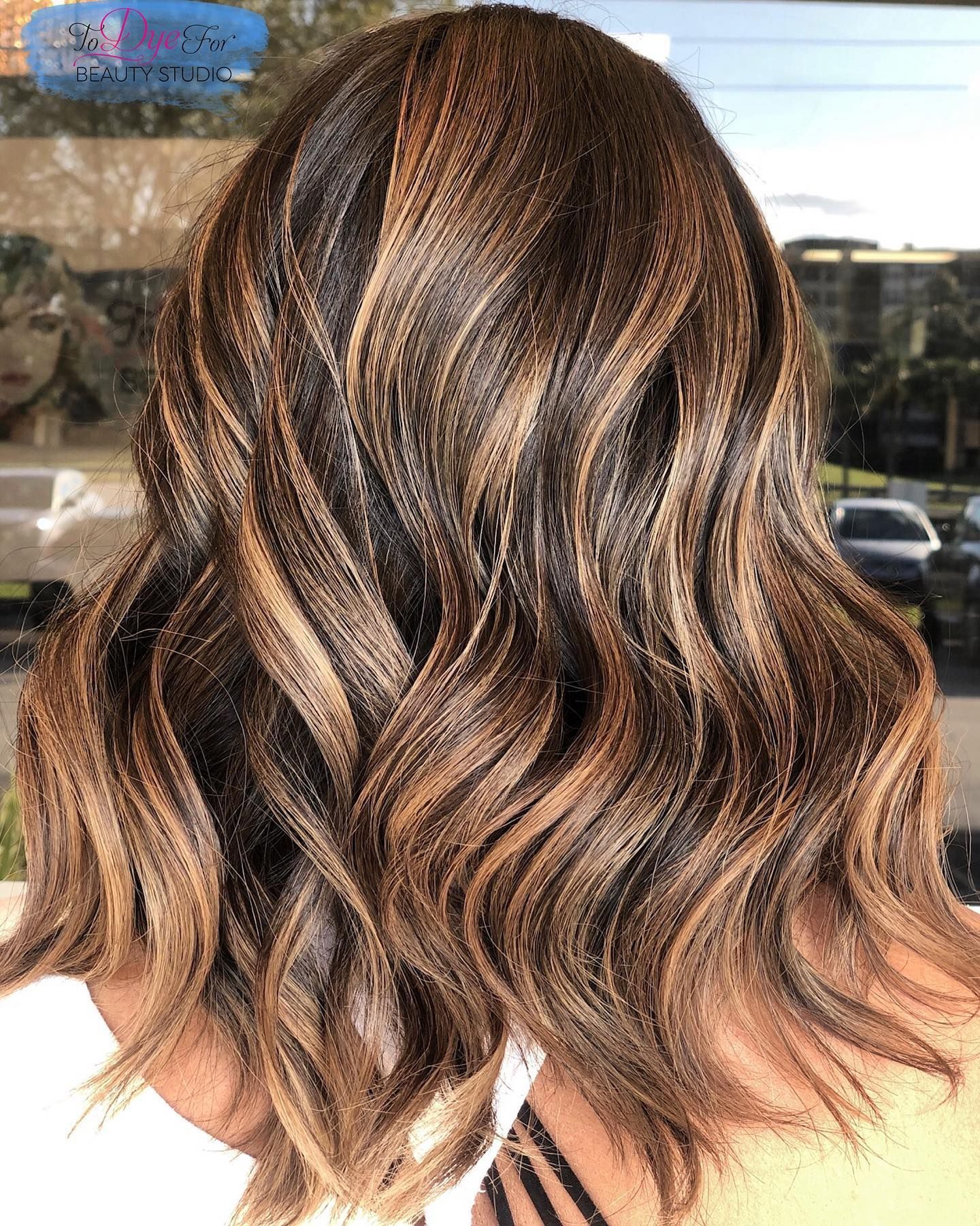 A woman with long wavy brown hair is standing in front of a window.