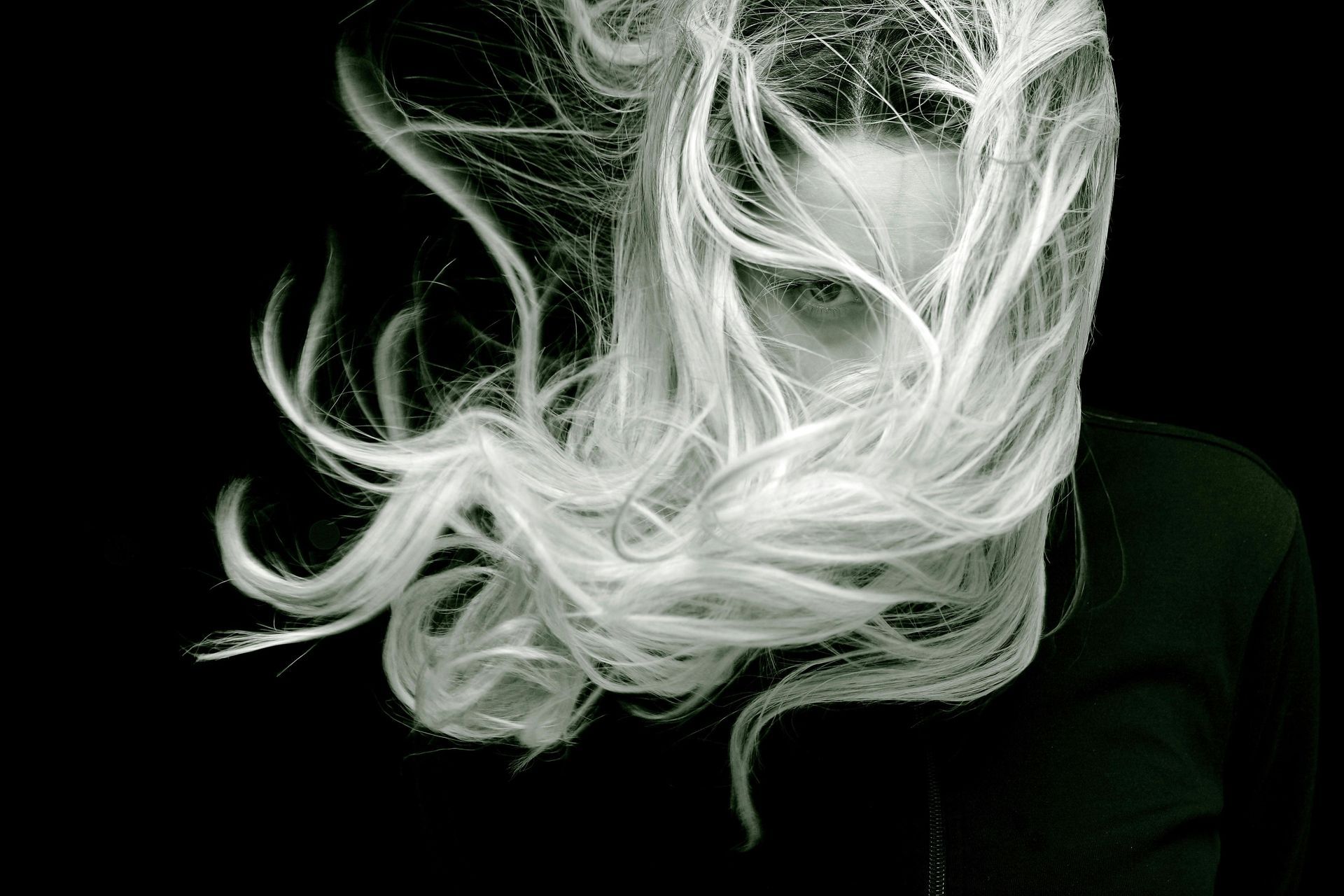 Blonde-haired person with hair obscuring face against a black background; only eyes visible.