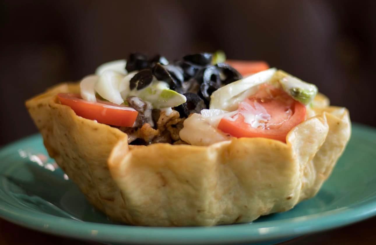 Taco salad in a crispy tortilla bowl, topped with tomatoes, olives, and onions, on a teal plate.