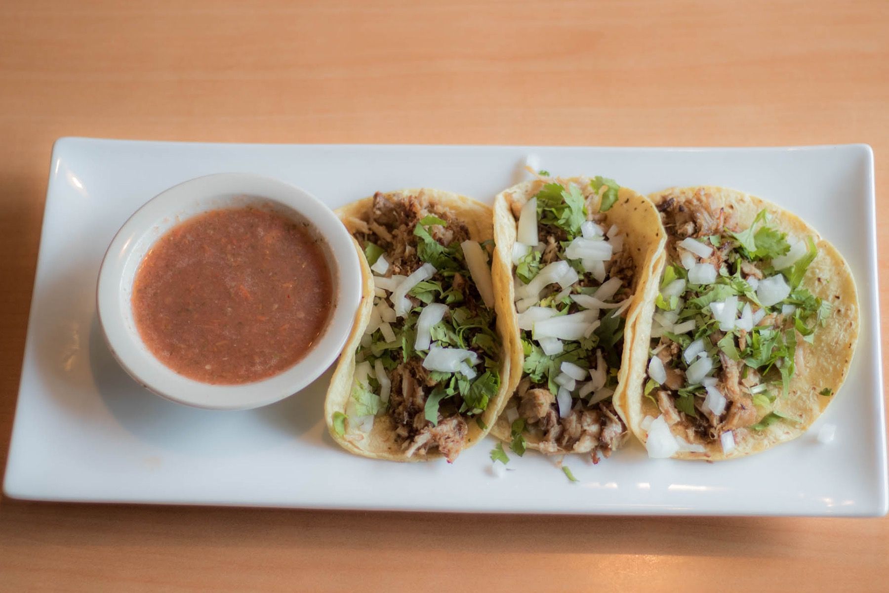 Three tacos on a rectangular white plate, with a small bowl of salsa.