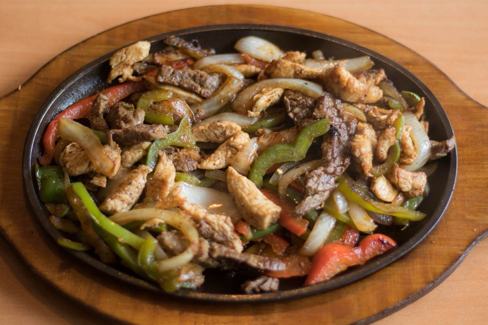 Sizzling fajitas with chicken, steak, peppers, and onions on a hot cast iron skillet, wooden base.