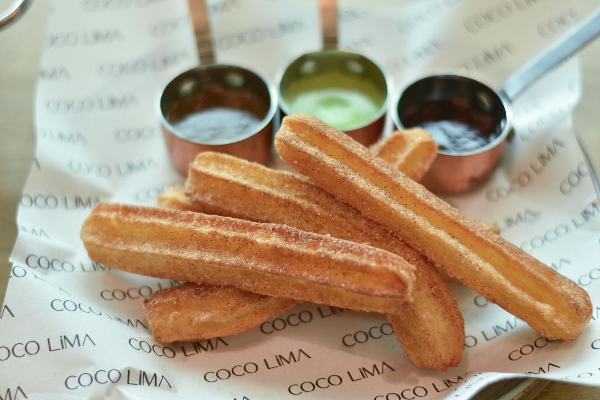 Churros with three dipping sauces, served on paper with a restaurant logo.
