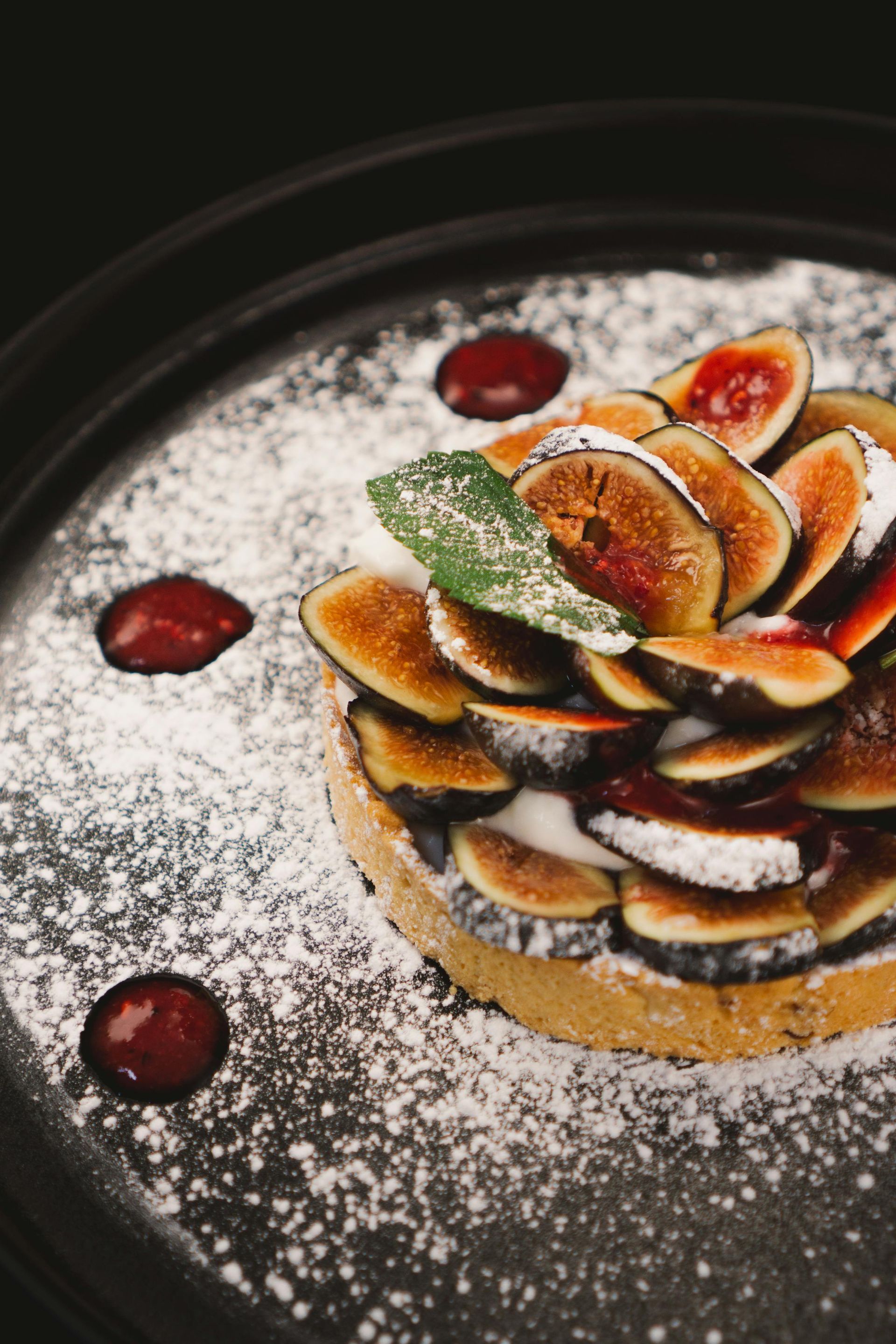 Fig tart on a black plate, dusted with powdered sugar, garnished with sauce and mint.