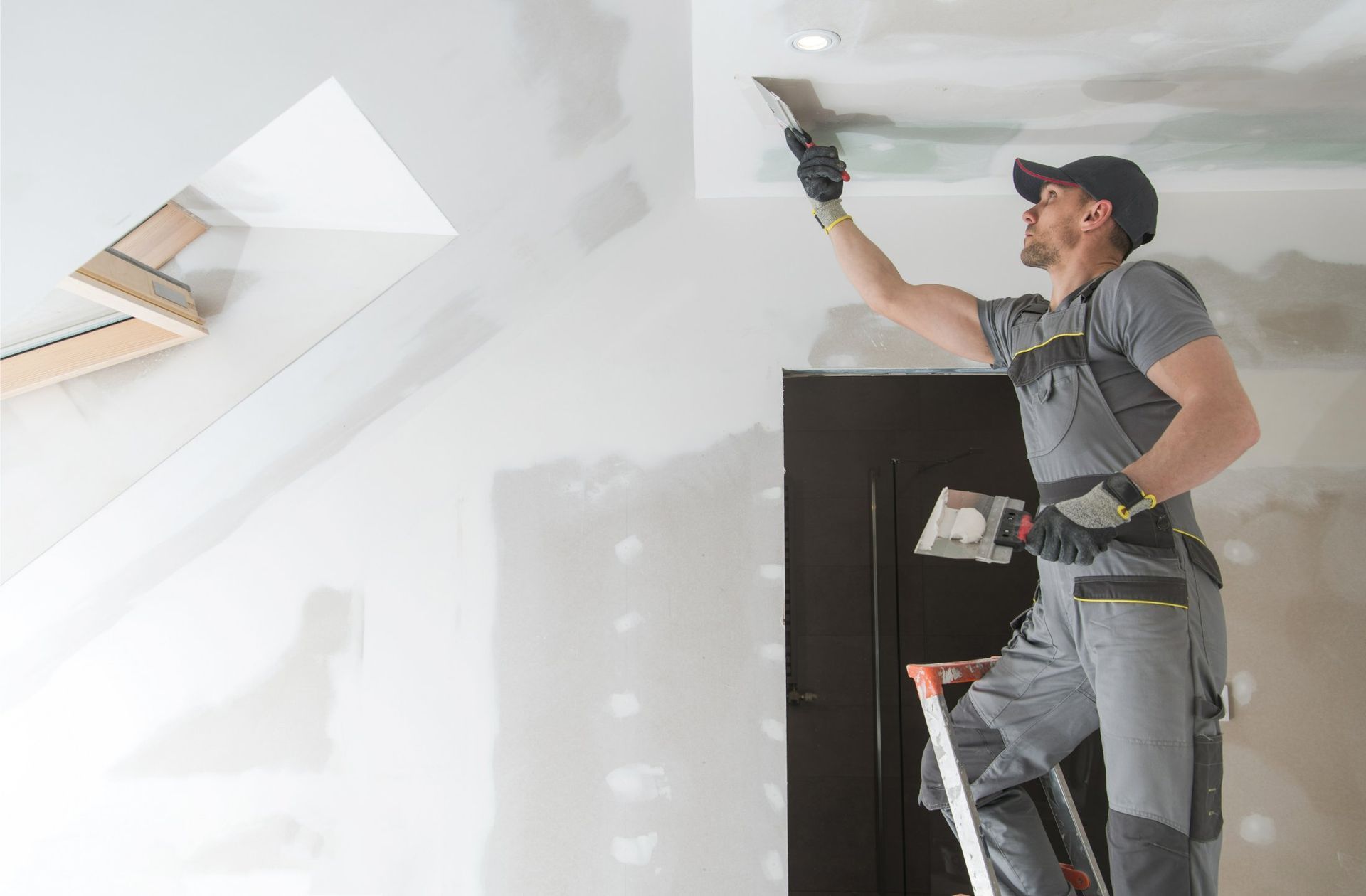 Person on a ladder, applying drywall compound to a ceiling with a trowel.