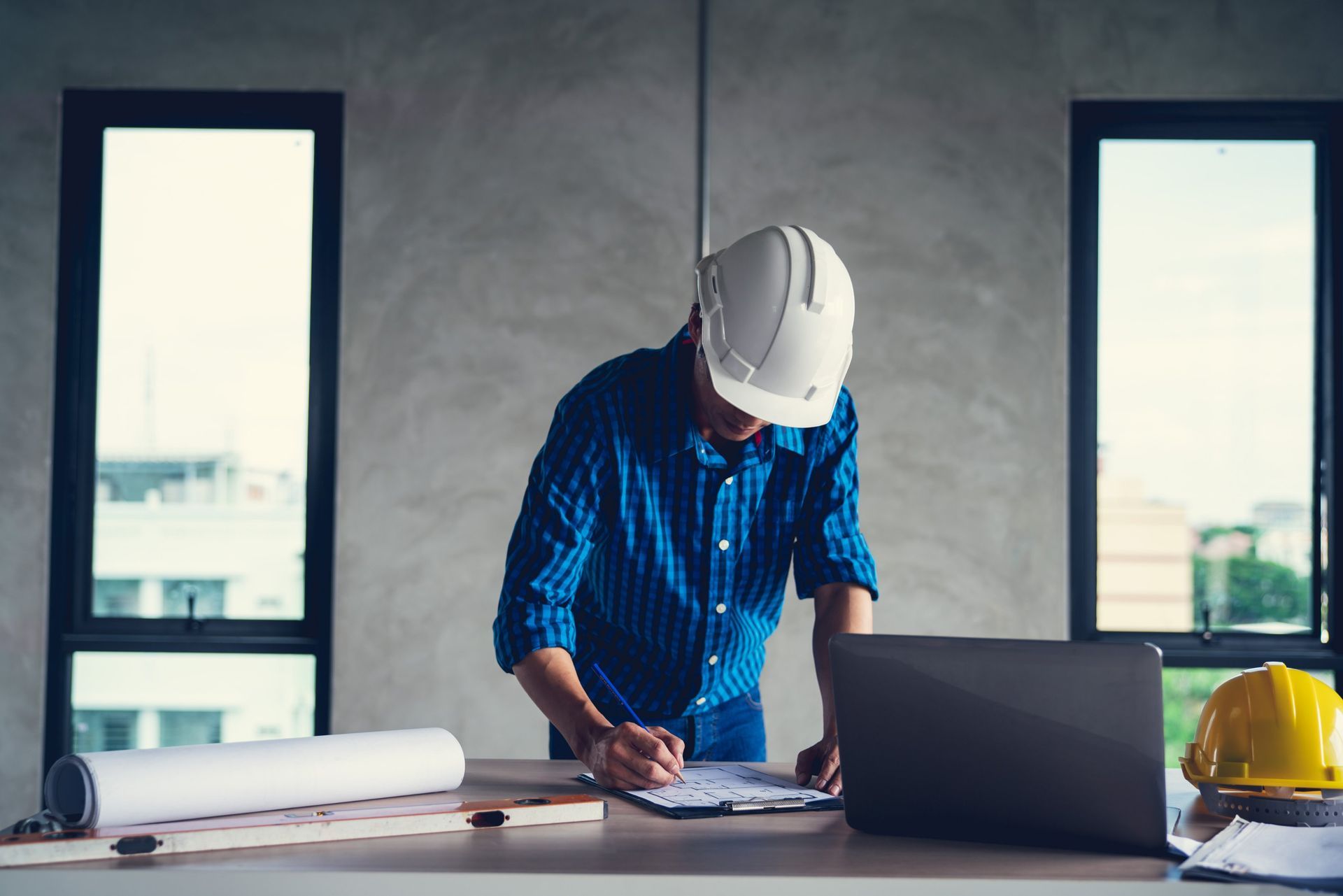 Construction worker in a blue shirt and white hard hat, working on plans at a desk with a laptop.