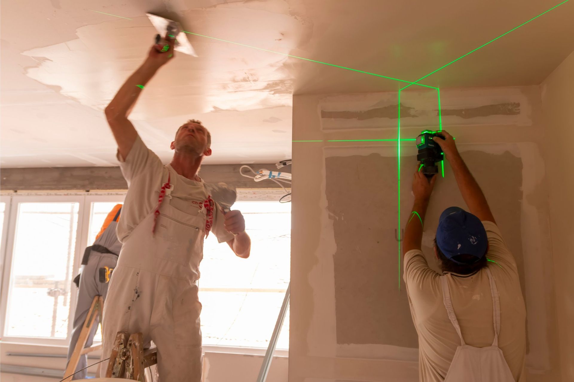 Two workers plastering a ceiling. One on a ladder, the other using a laser level, green lines visible.