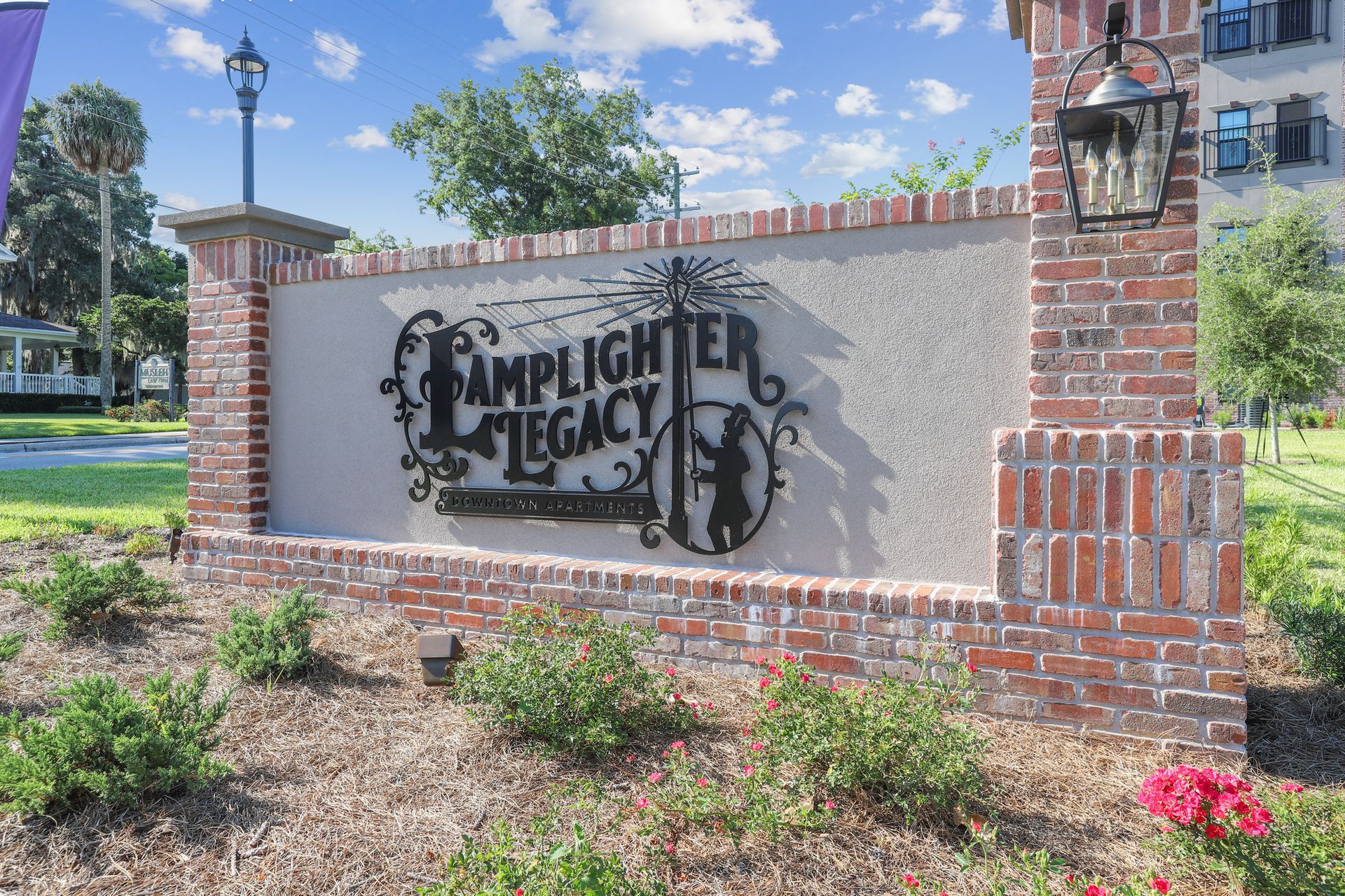 Sign for Lamplighter Legacy apartments; brick and stucco structure, ornate metal sign, greenery.