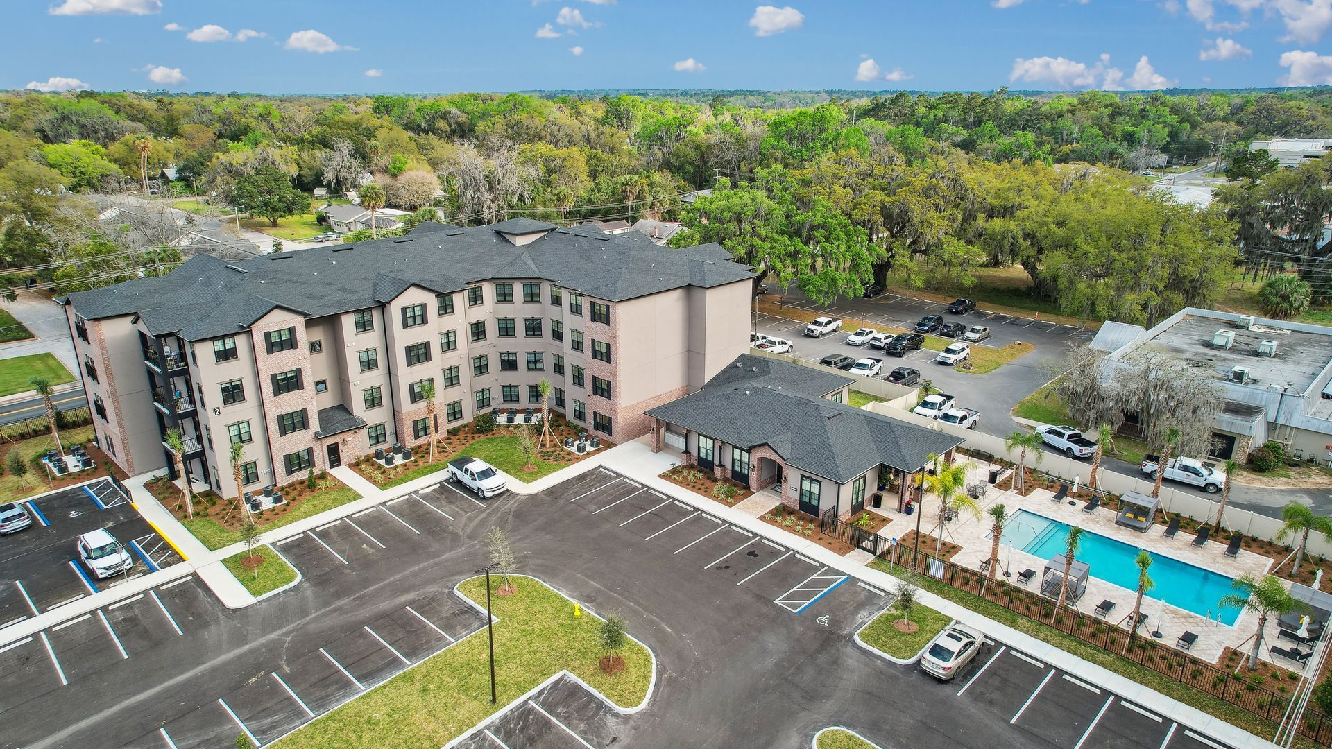 Apartment building with pool and parking lot, surrounded by trees, blue sky.