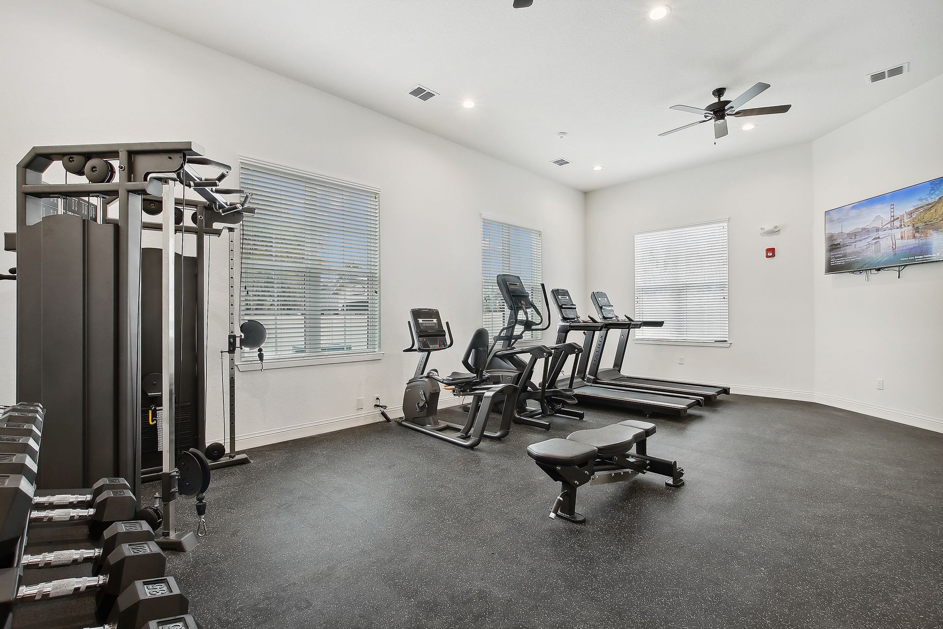 Gym with weight machine, exercise bikes, treadmills, bench, and dumbbells; rubber floor and white walls.