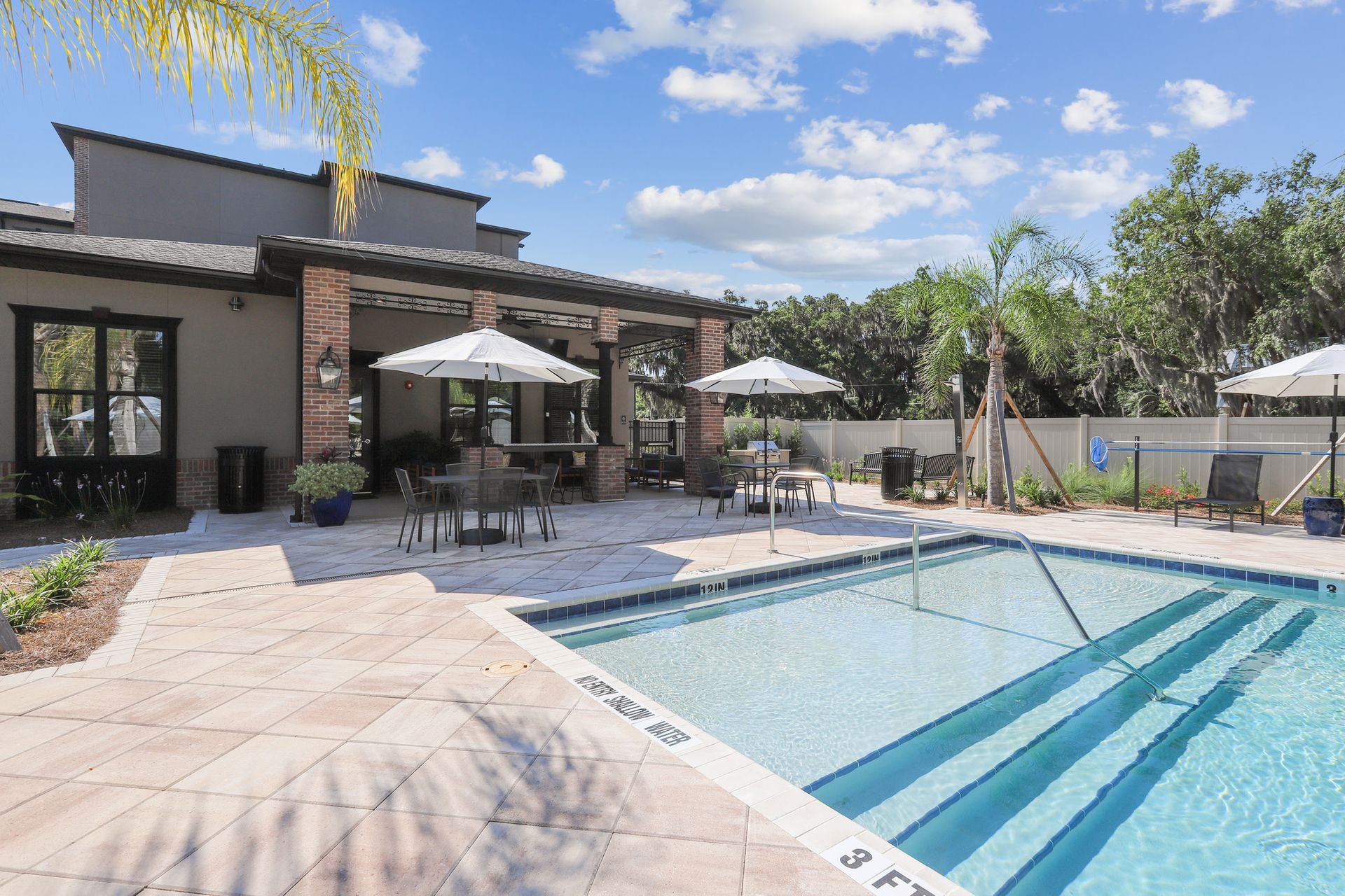 Pool with steps, tables, umbrellas, and a building in the background on a sunny day.