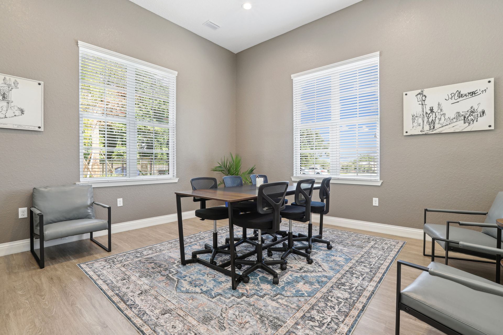 A modern meeting room with a table, chairs, rug, and two large windows. Gray walls and light wood floors.