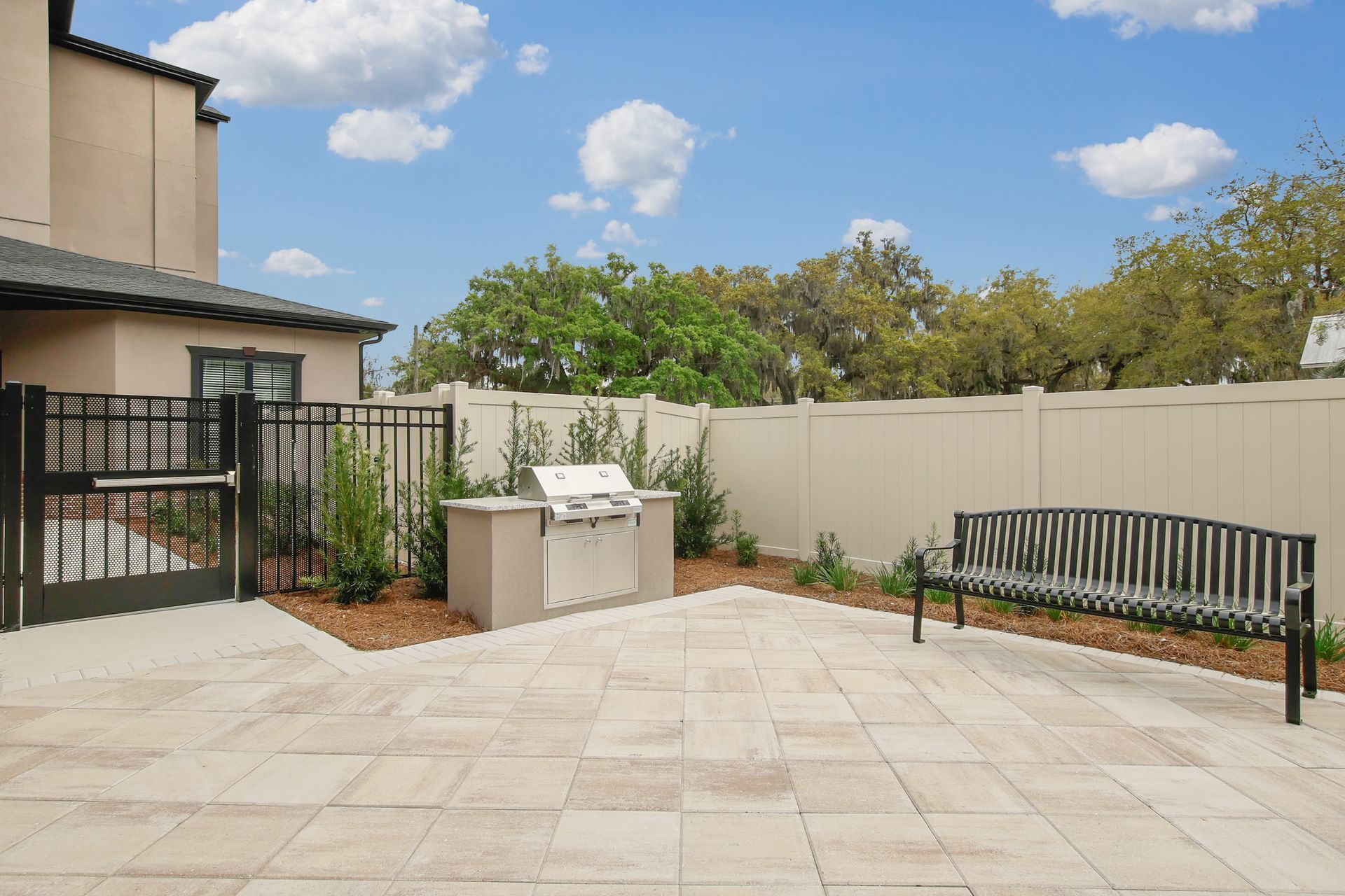 Outdoor patio with grill, bench, tan pavers, and beige fence under a blue sky.
