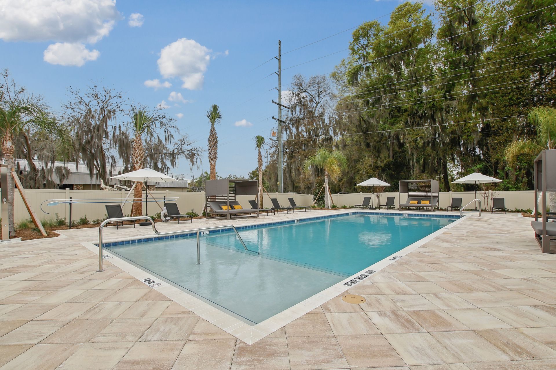 Swimming pool surrounded by lounge chairs and umbrellas. Sunny day, trees in the background.