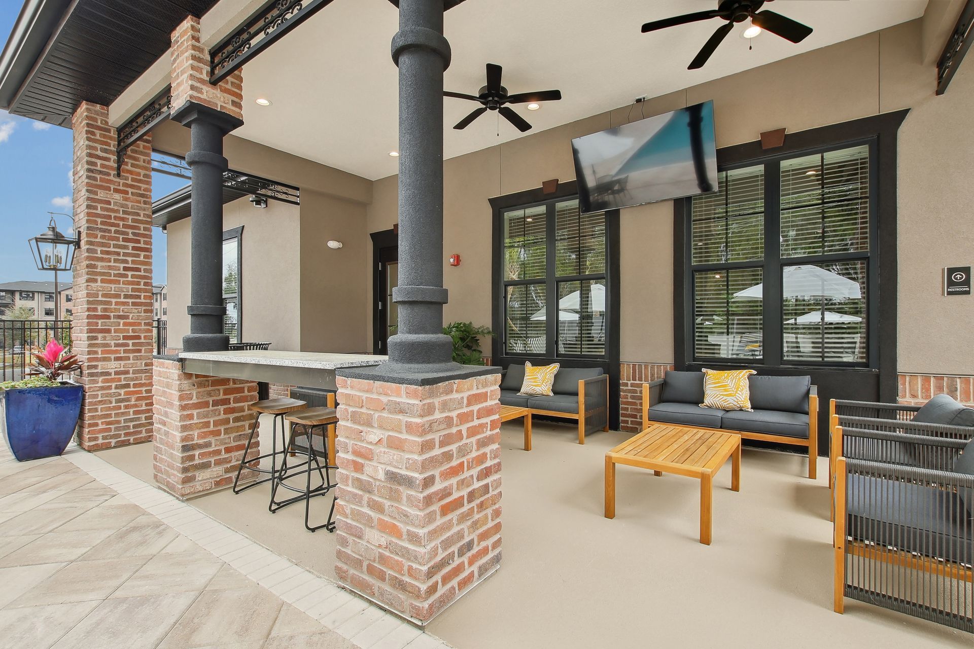Outdoor patio with seating, TV, brick columns. Gray couches with yellow pillows.