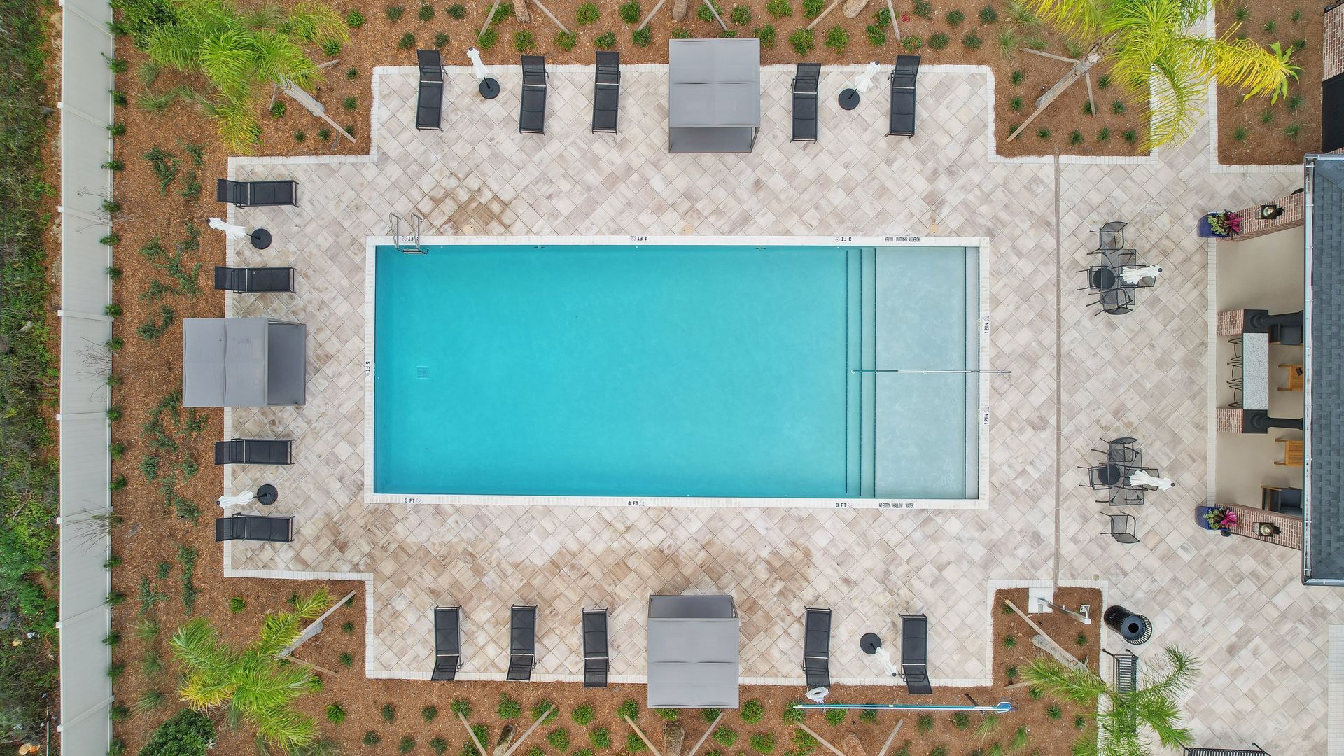 Overhead view of a rectangular pool surrounded by lounge chairs, gazebos, and a building; beige and blue colors.