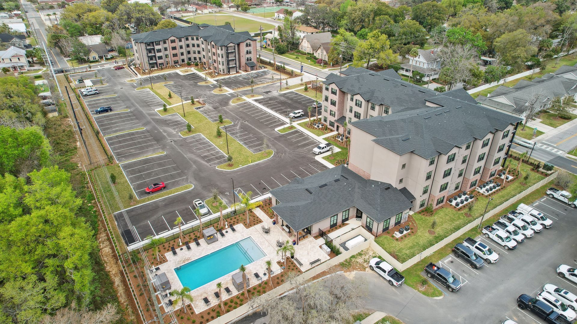 Aerial view: apartment complex with pool, parking, and landscaping. Buildings are beige with dark roofs.