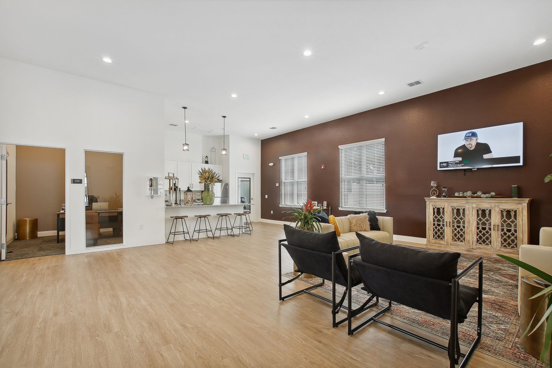 Modern living room with seating area, kitchen, and a mounted TV. Light wood floors and dark accent wall.