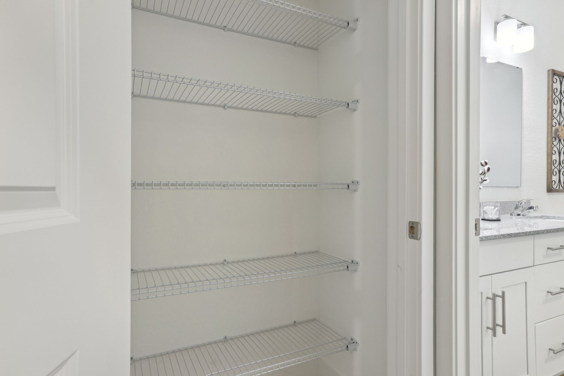 White closet with wire shelves, open door to a bathroom with a vanity and mirror.