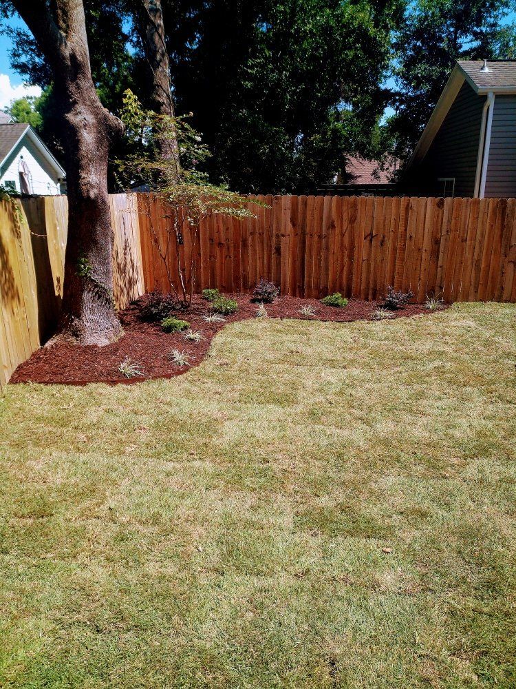 Backyard with a wooden fence, mulch beds, a large tree, and a grassy lawn under bright sunlight