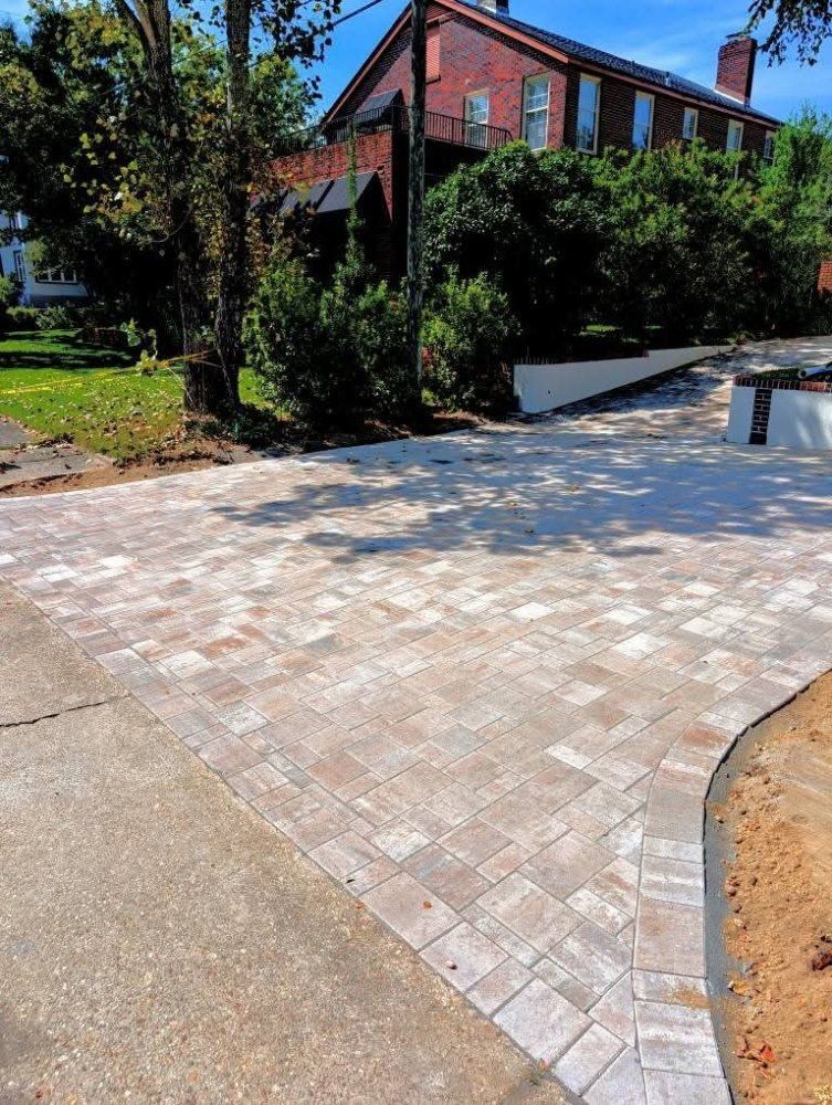 Brick-paved driveway beside a red brick house, bordered by trees and greenery on a sunny day