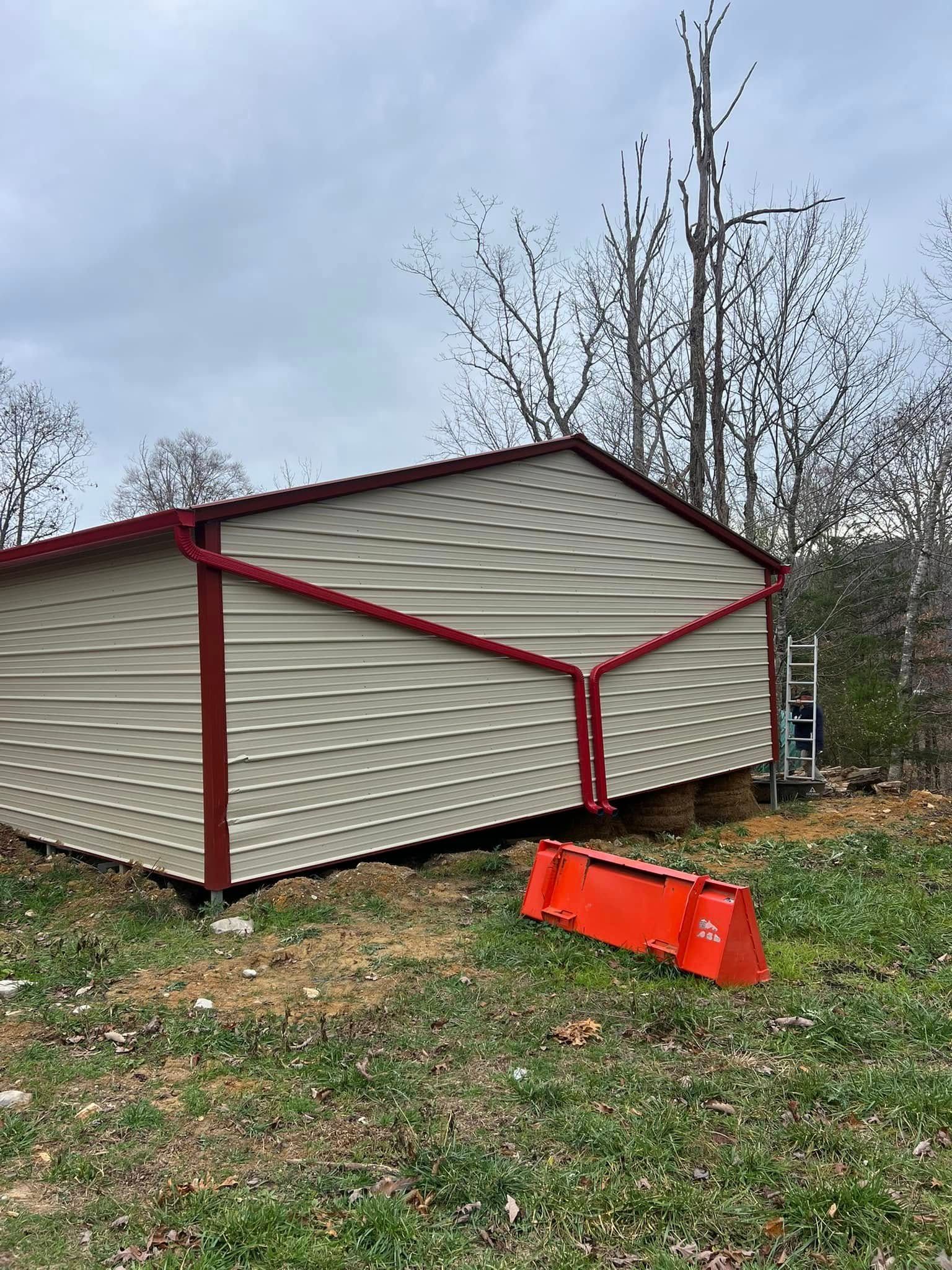 A beige metal shed with red trim stands in a grassy field under a cloudy sky, with a bright orange piece of equipment nearby.