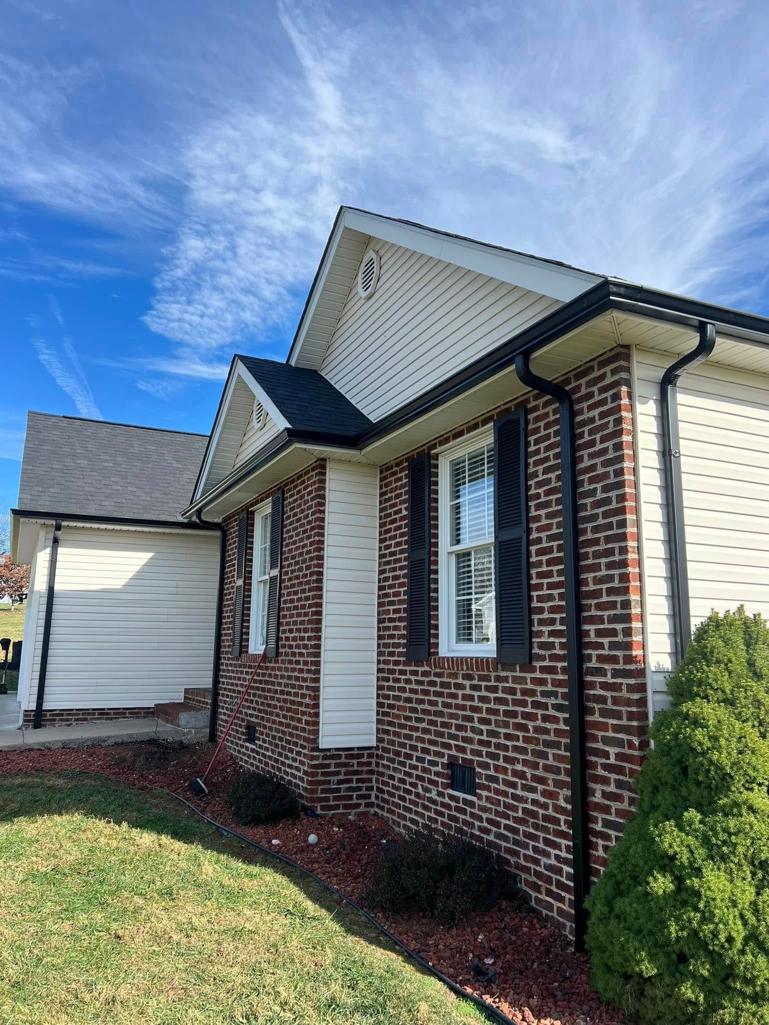A low-angle view of a brick house with light siding, dark shutters, and a shingled roof under a bright blue sky.