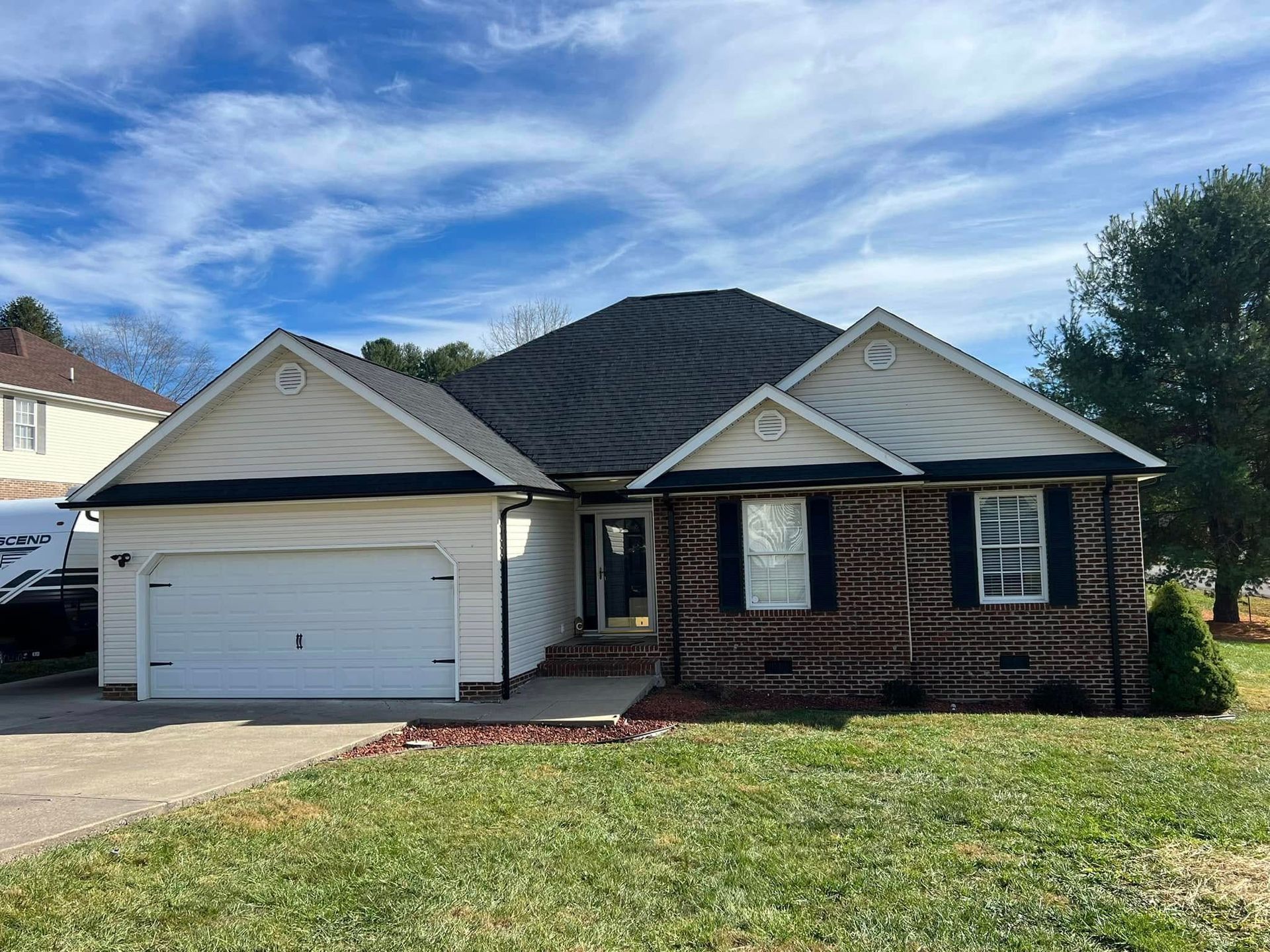 A one-story suburban home with a two-car garage, brick and light siding, and a dark roof under a blue, cloudy sky.