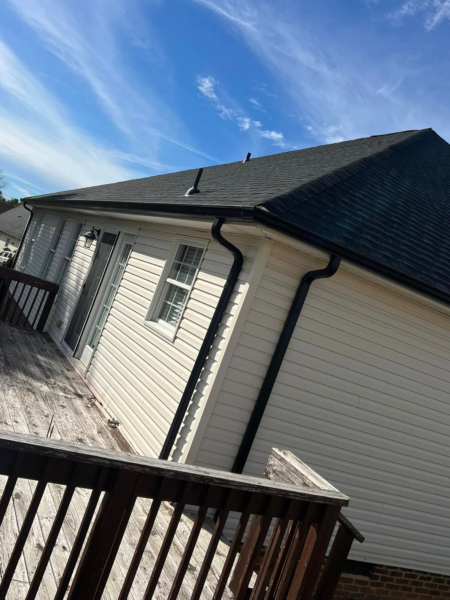 A side view of a cream-colored house with black gutters and a dark roof under a bright blue sky with thin clouds.