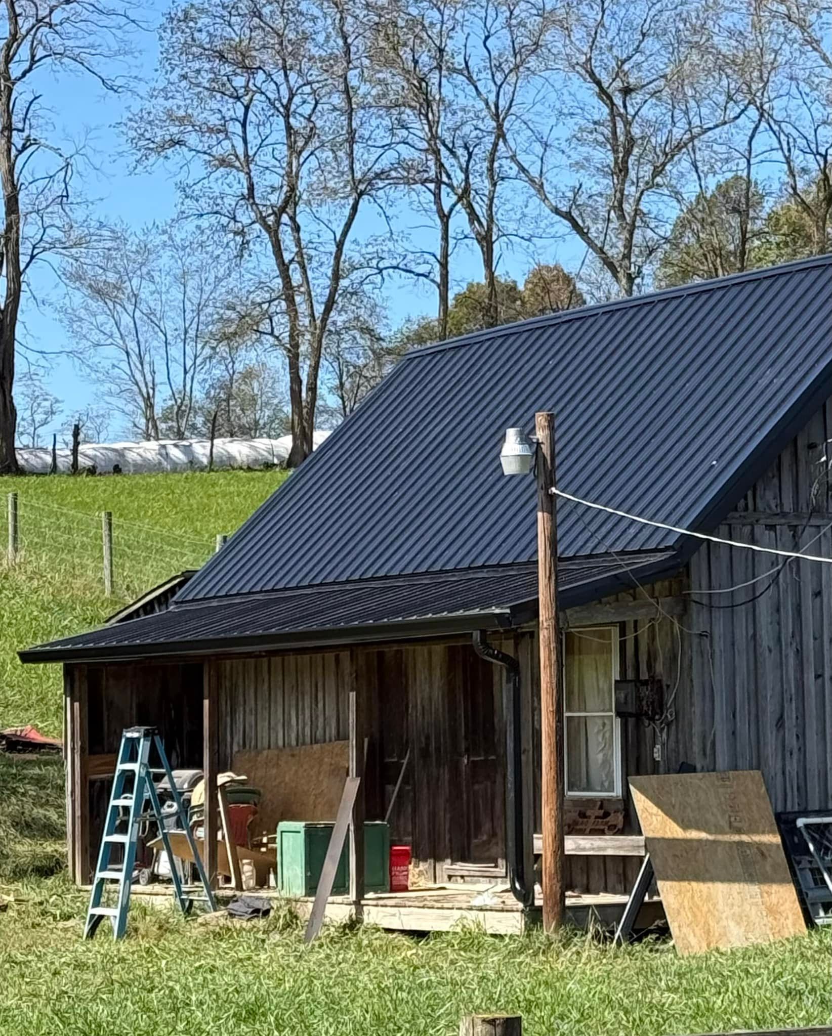 A rustic wooden barn with a dark metal roof sits on a grassy hill under a blue sky, with a ladder leaning against it.