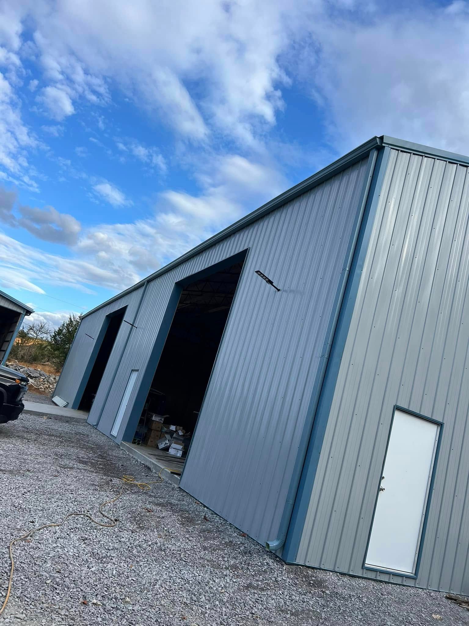 A large, modern metal industrial warehouse with open bay doors and a white pedestrian door against a cloudy blue sky.