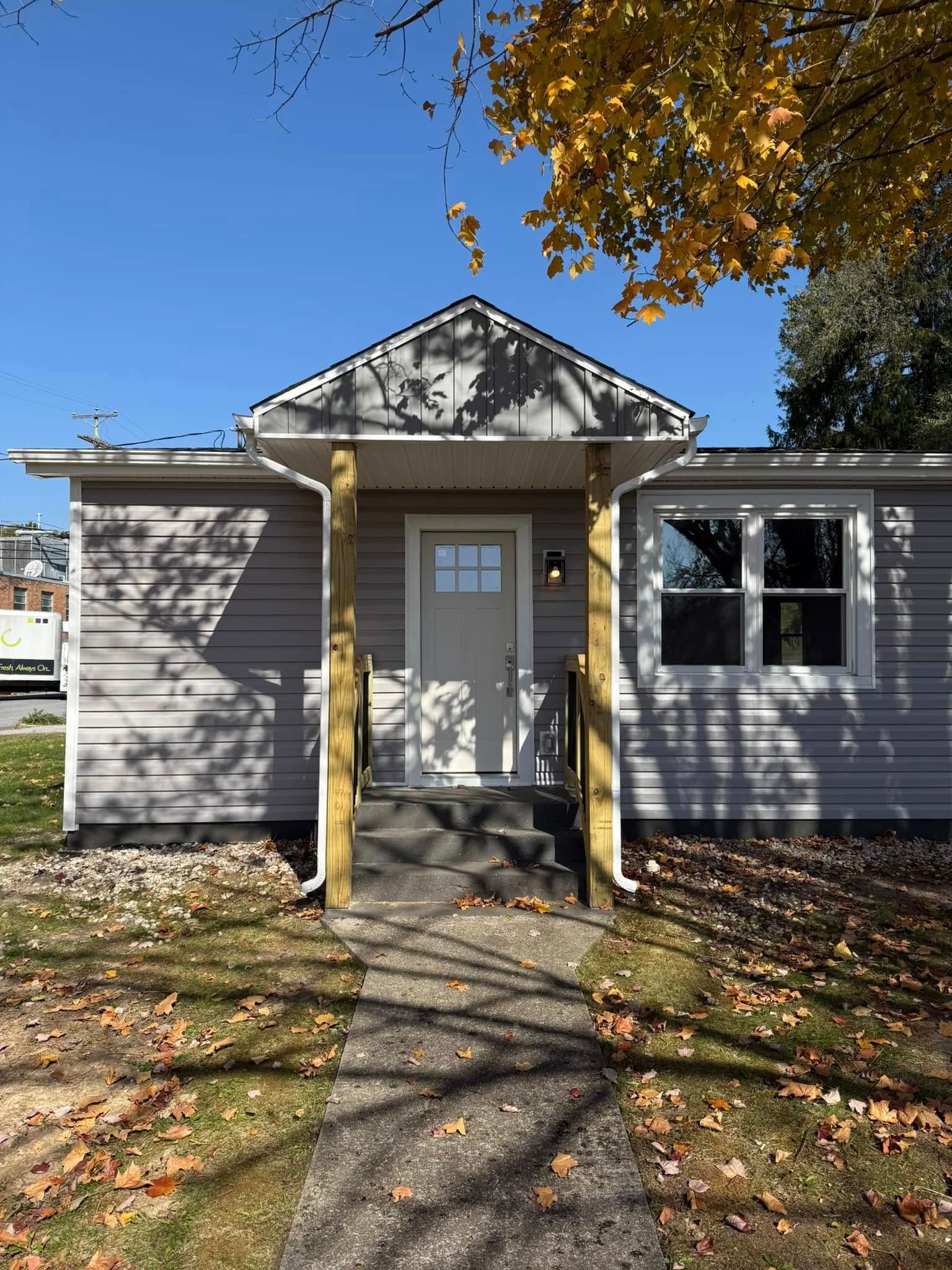 A gray, single-story house front with a covered entrance porch, wooden posts, and a sidewalk leading to the front door.