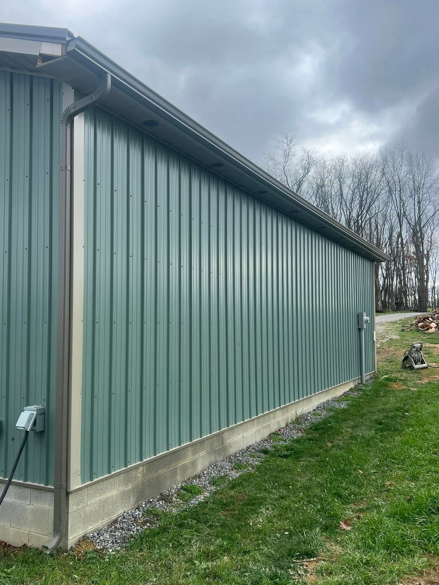 A side view of a green corrugated metal building with a concrete foundation on a grassy, sloped hill.