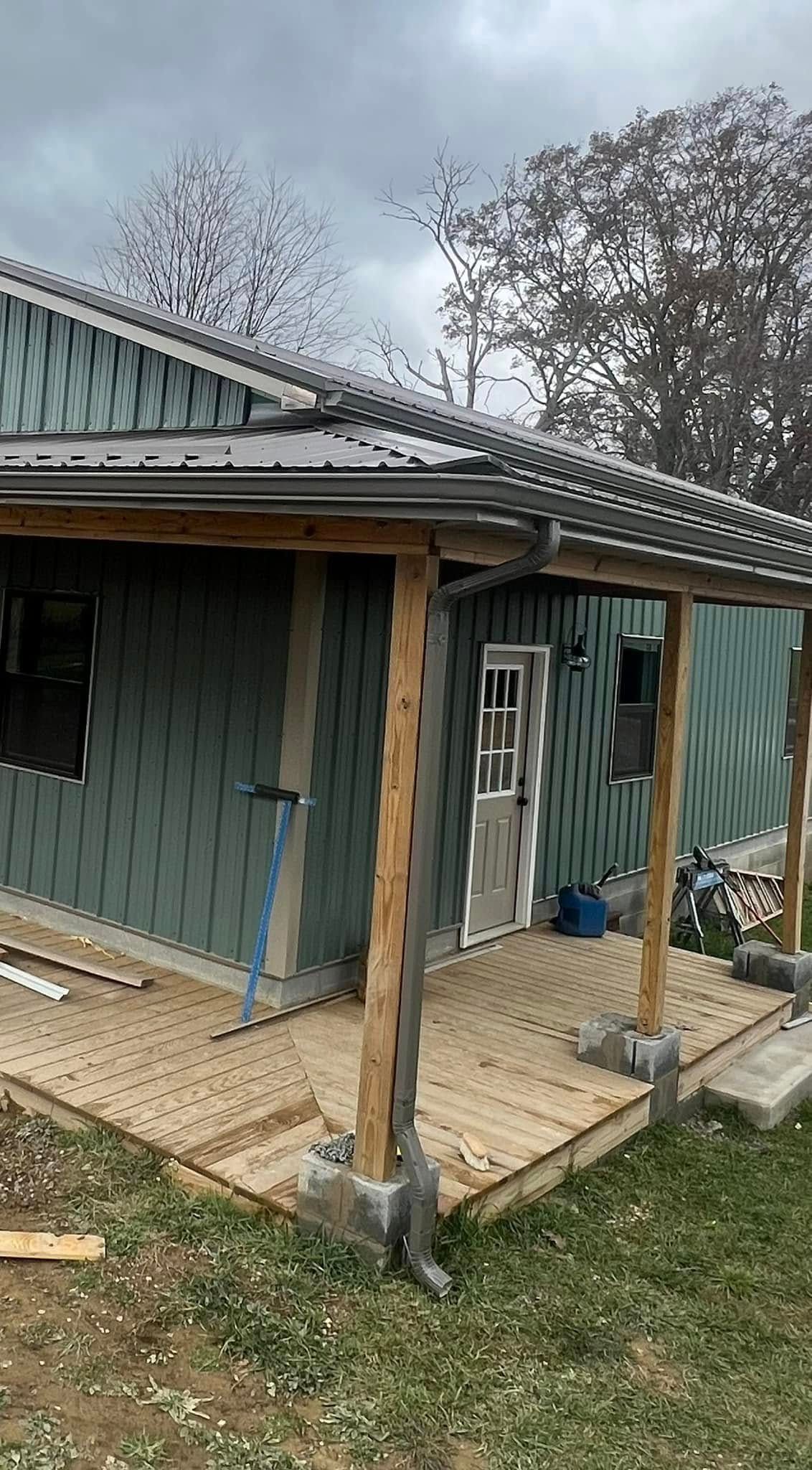 A house under construction with green siding, a metal roof, and a wooden porch supported by posts on concrete blocks.