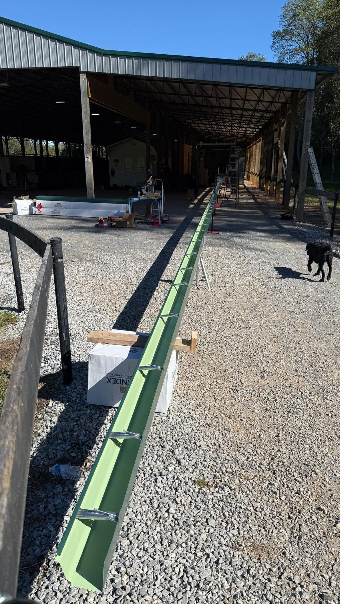 A long green metal livestock feeder trough sits on a gravel driveway in front of a covered barn, with a dog walking nearby.