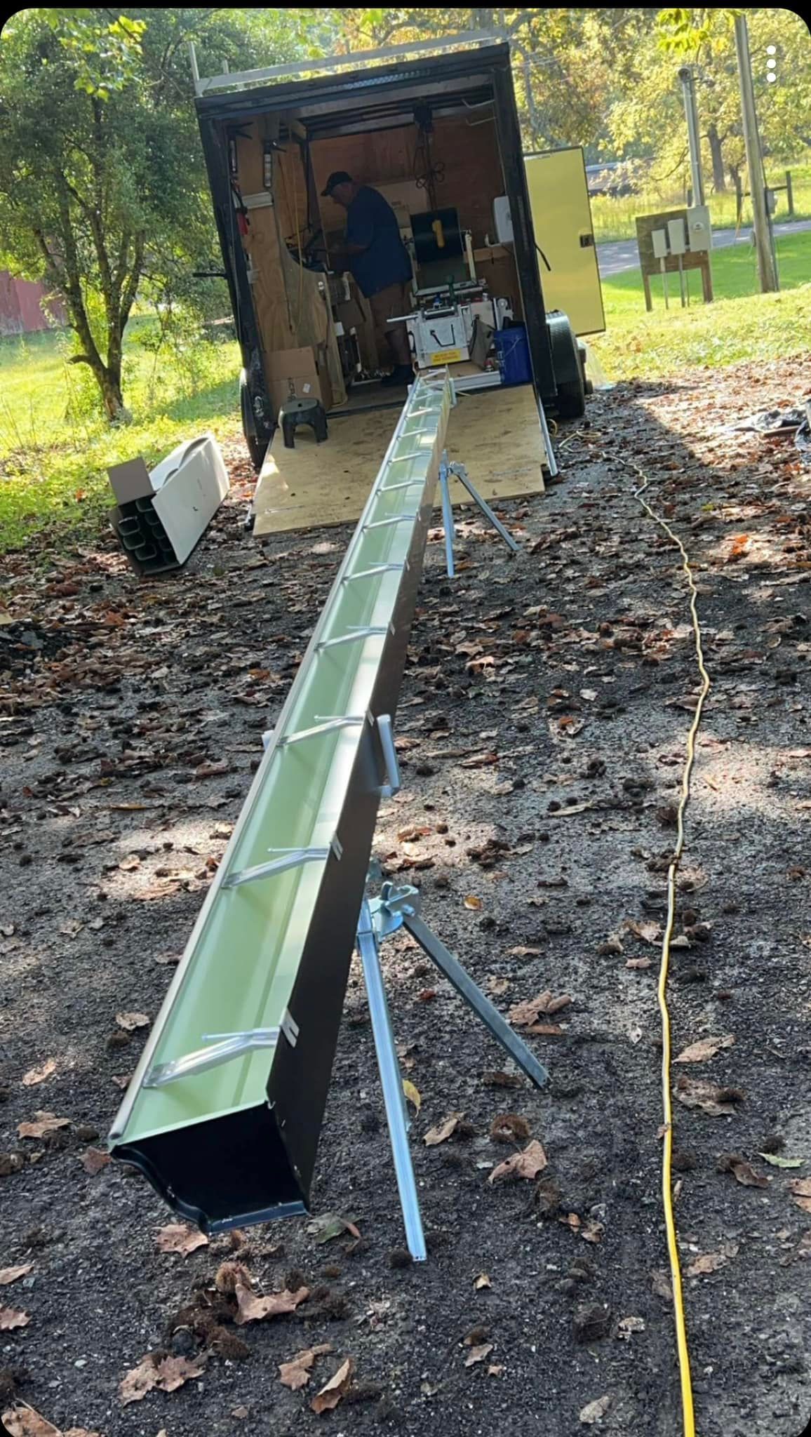 A long, green seamless gutter rests on a metal stand extended from a mobile workshop trailer on a gravel lot.
