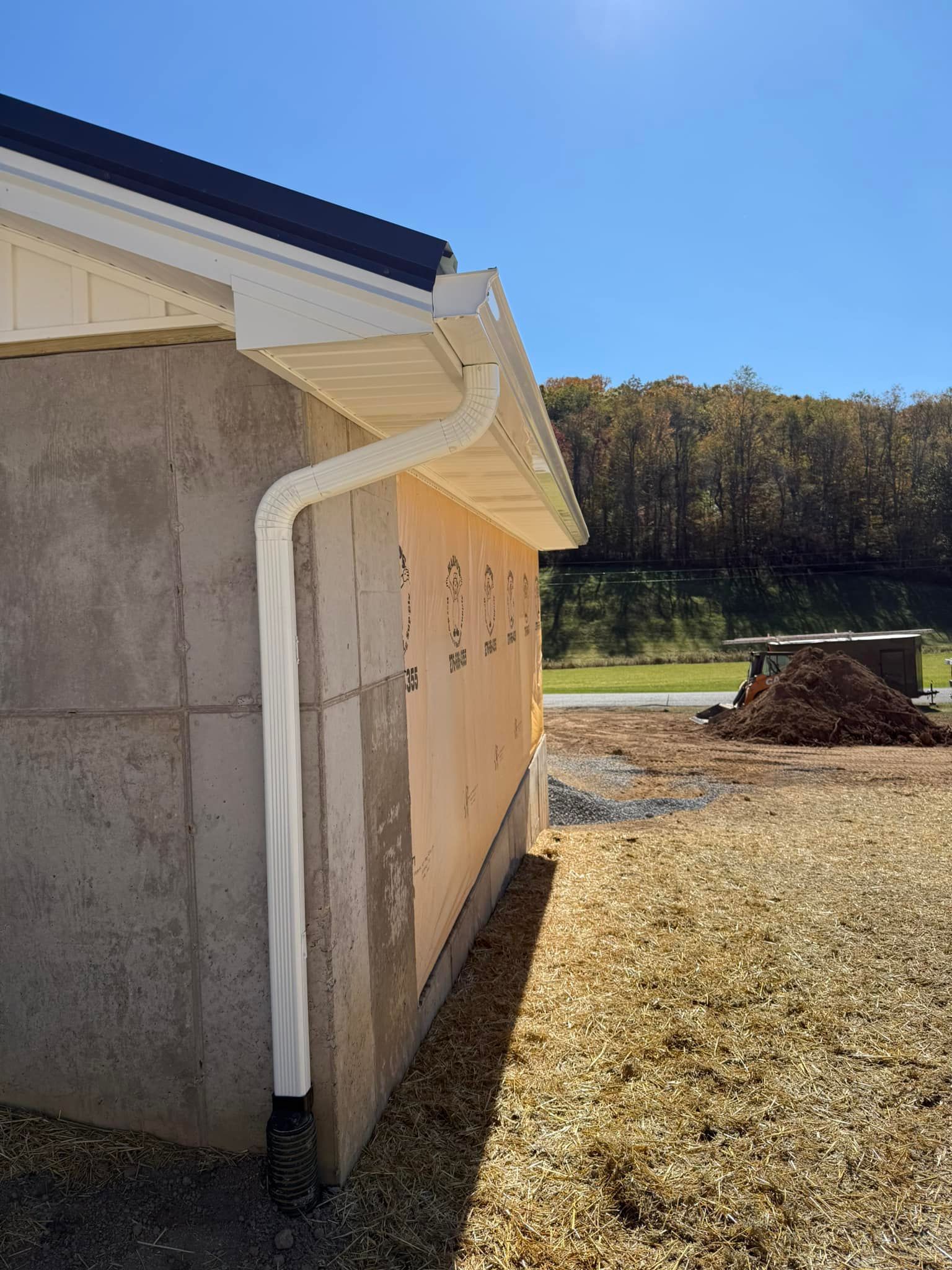 White downspout installed on the corner of a building exterior with light-colored siding and a forest background.