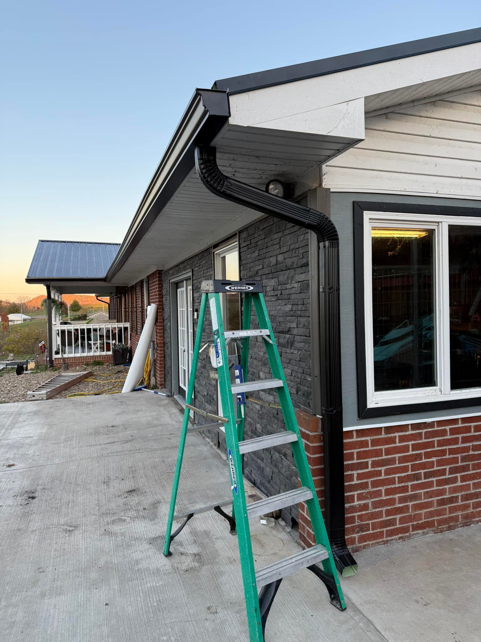 A black gutter downspout installation on a house exterior next to a green step ladder and a concrete patio.
