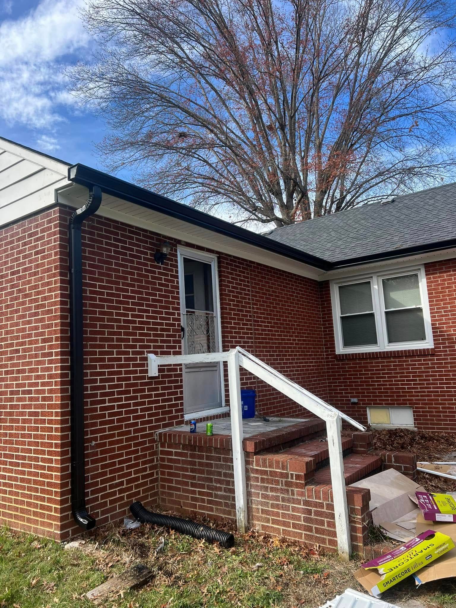 Red brick exterior of a house with a small porch, white railing, black gutters, and a bare tree against a blue sky.