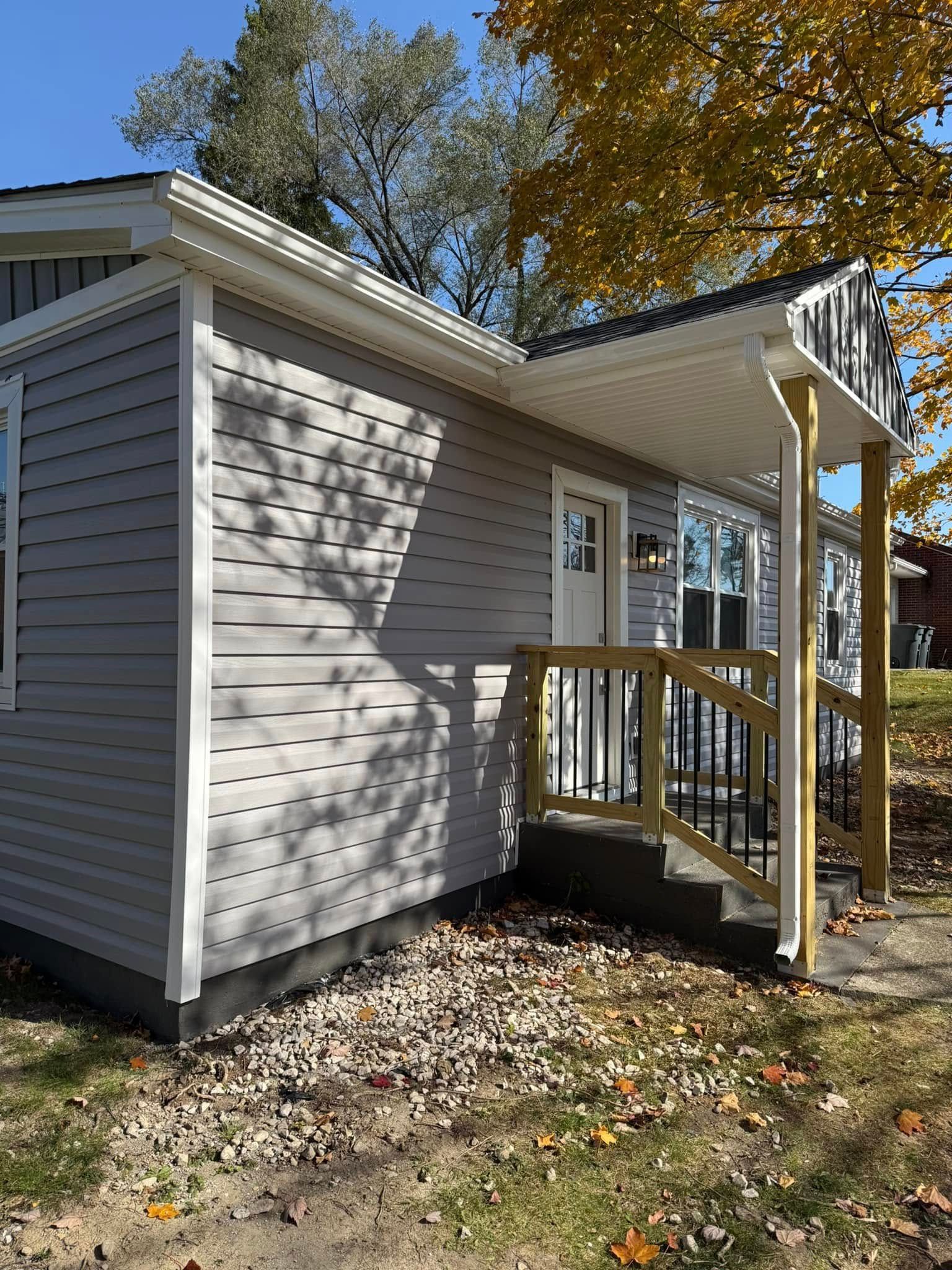 Exterior view of a gray vinyl-sided house with a small front porch, wooden railing, and stairs, surrounded by fall leaves.