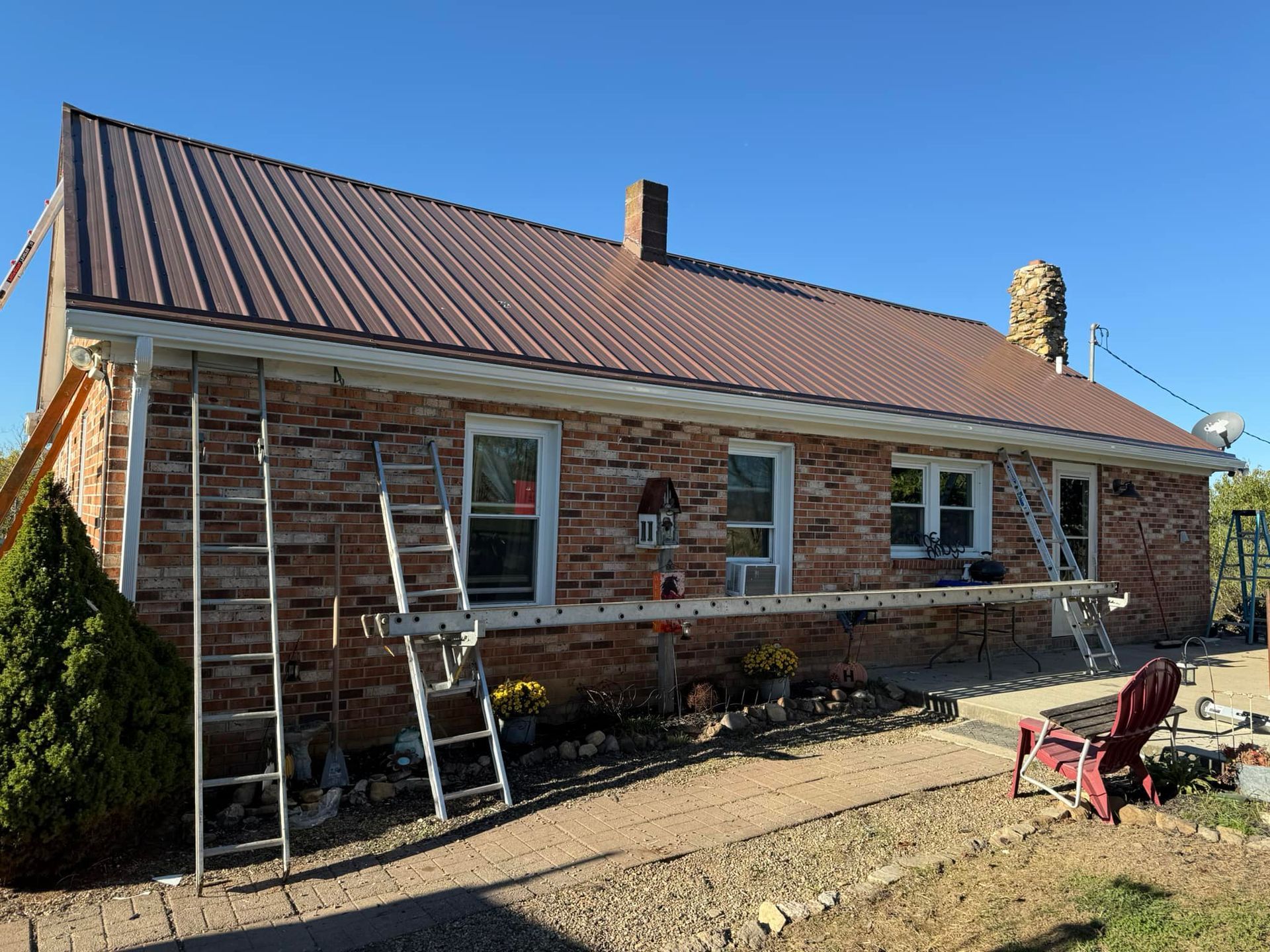 A brick house with a brown metal roof sits under a clear blue sky, with construction ladders set up along the wall.