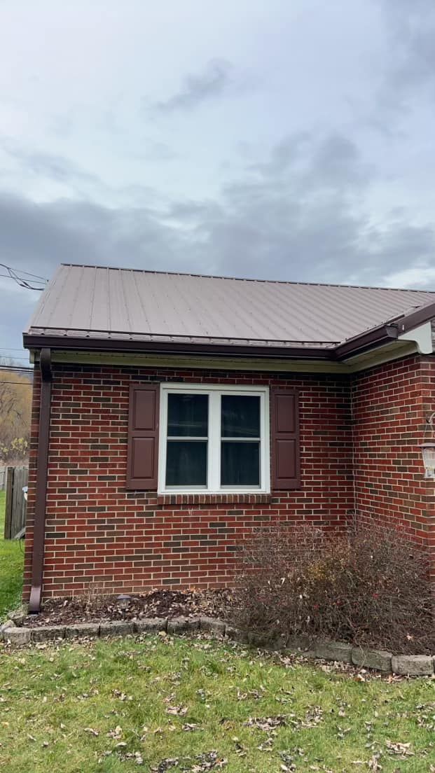A brick house corner with a brown metal roof, white-framed window, dark shutters, and a downspout against a cloudy sky.