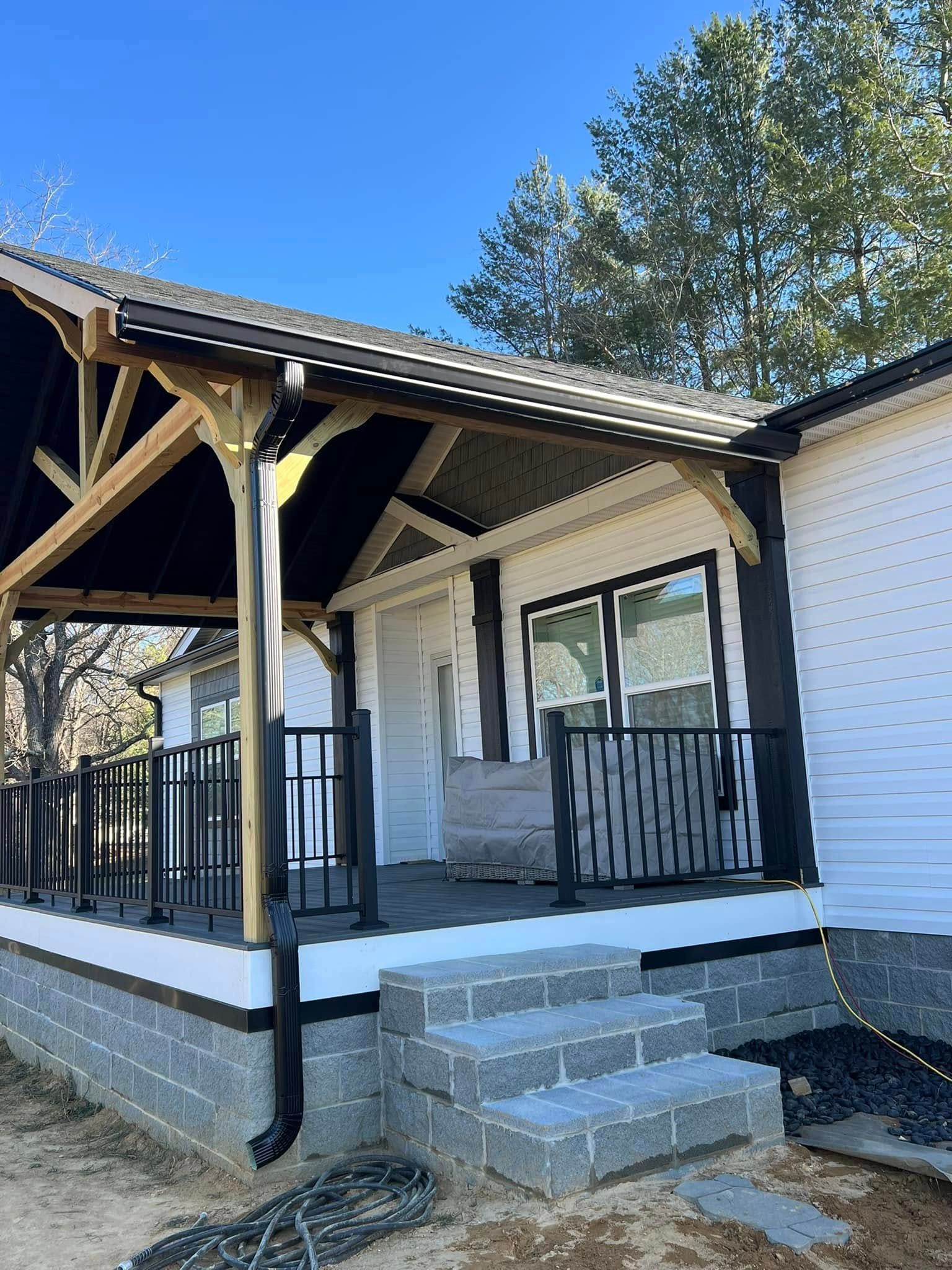 A white house with a covered wooden porch, black railings, and gray block steps against a blue sky.
