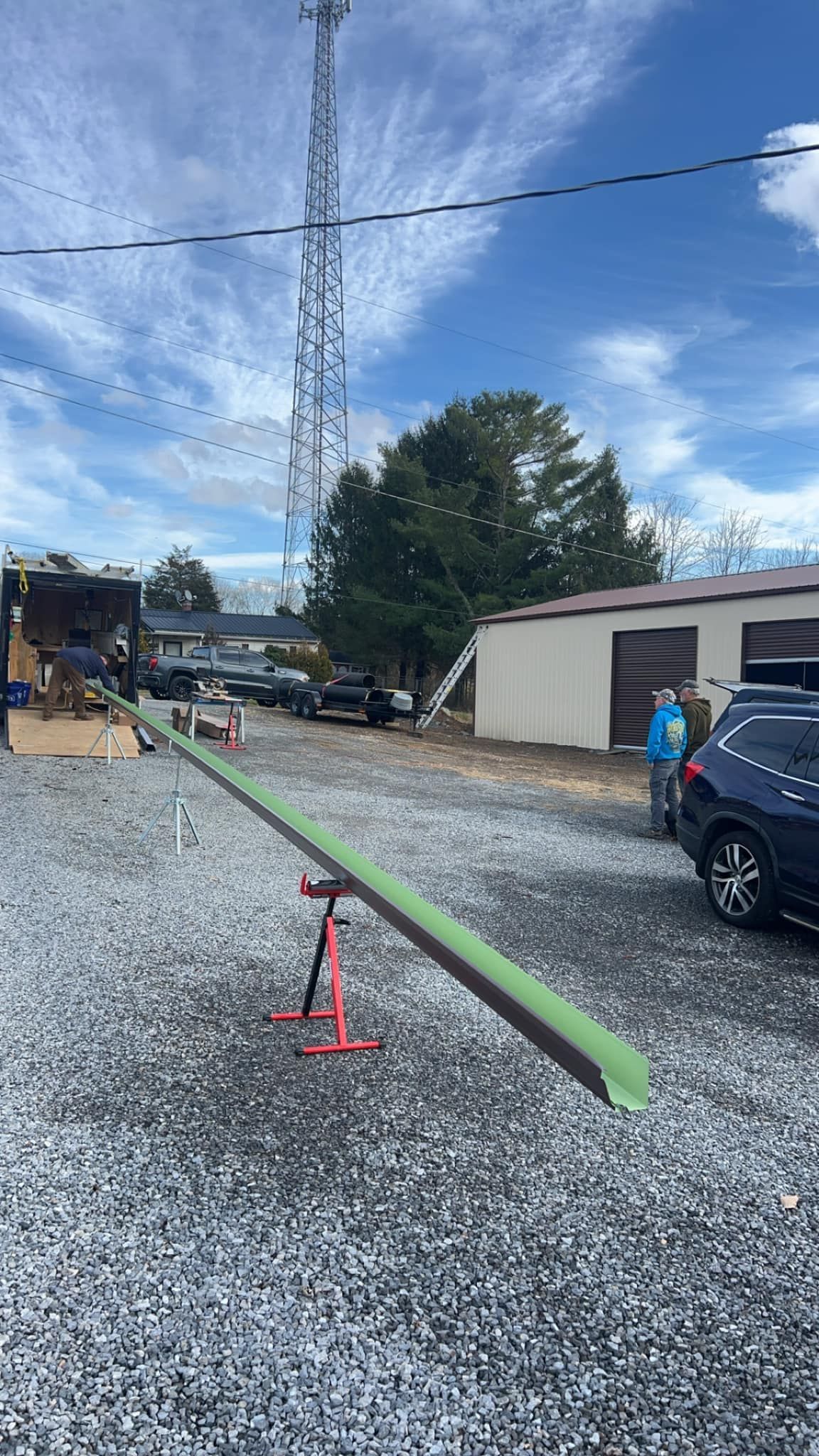 A long, light green gutter section rests on a red stand in a gravel lot with a radio tower and buildings in the background.